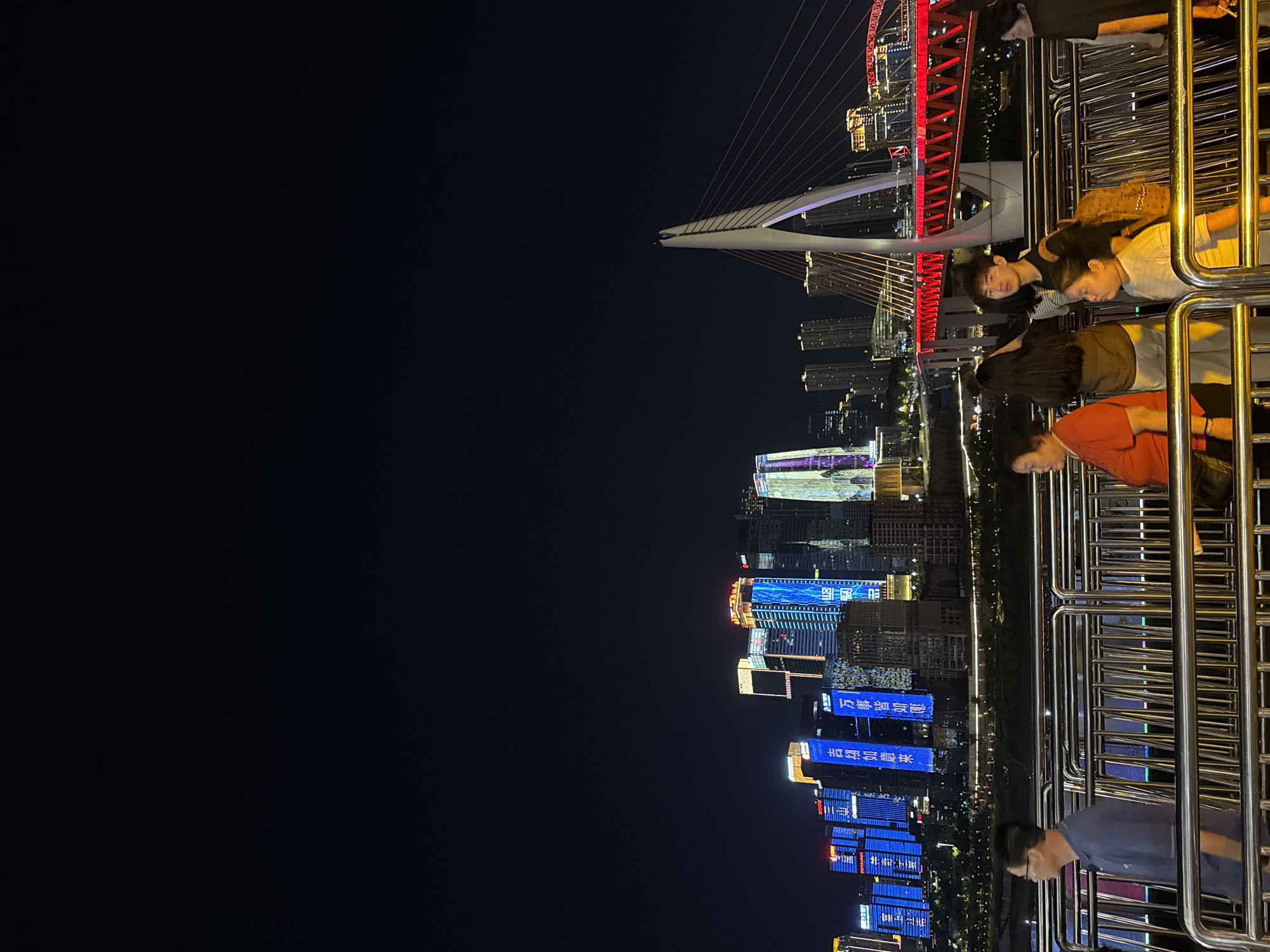 At night, a group of people are walking on a pedestrian bridge with a railing in the foreground. The people include a man in a blue polo shirt, a woman in an orange shirt, a woman with long black hair, a young man with a backpack, a young girl in a white shirt, and a woman in a black shirt. In the background, a modern cityscape is illuminated with bright lights. Several tall buildings with colorful neon signs are visible, including a building with blue vertical signage. A cable-stayed bridge with red lighting stretches across a body of water. The scene is located in Yuzhong District, China, a district known for its vibrant nightlife and modern architecture along the Yangtze River.