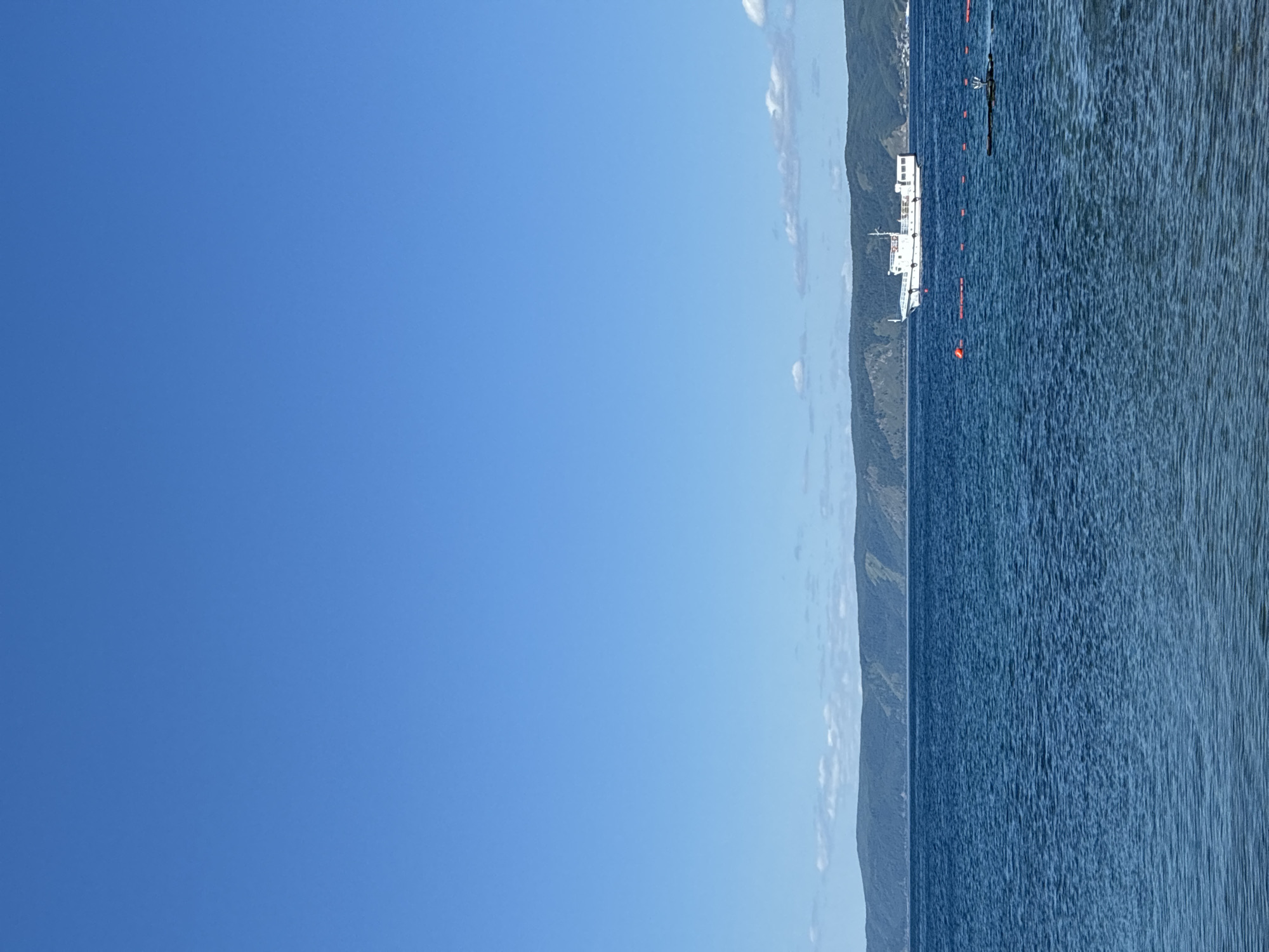 A white passenger boat with multiple windows and a raised deck is cruising on a large, calm, blue body of water under a clear, bright blue sky. In the distance, a long, low mountain range covered in dark green trees stretches across the horizon, with hints of lighter vegetation and some structures visible on the far right. Several red buoys are floating on the water in the foreground, and a dark, rectangular object with a white pole and a small flag is partially submerged on the right side of the frame. The water is rippled, indicating a gentle breeze. The scene depicts a tranquil day on the water, likely on Lake Baikal near Listvyanka, Russia, given the mountainous landscape typical of the region.