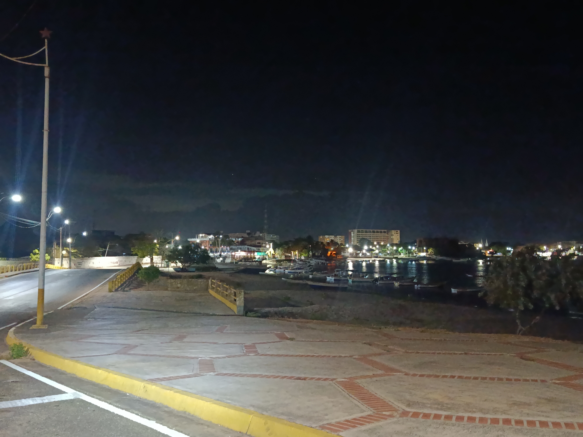 The image depicts a nighttime scene in Juan Griego, Venezuela. A paved area with red brick accents is in the foreground, bordered by a yellow curb. A road leads towards a harbor or bay, with several boats visible on the water. The background shows a cityscape illuminated by lights, including buildings, streets, and possibly some illuminated signs. A lamppost with a red star on top is situated on the left side of the image. The sky is dark and partially obscured by clouds.