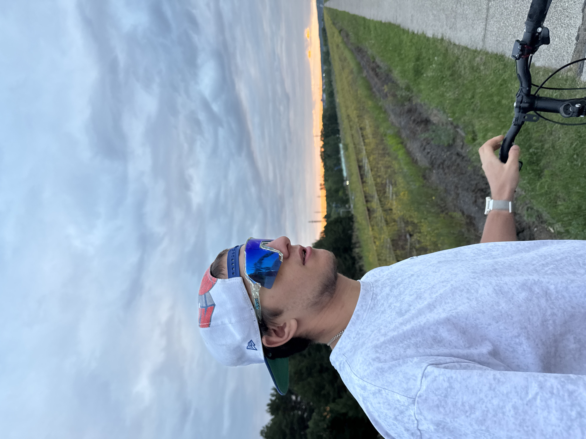A young man wearing a white New Era baseball cap and mirrored sunglasses is pictured from the chest up, looking to the right. He is wearing a light-colored, mottled t-shirt and a silver watch. He is holding onto the handlebars of a bicycle, suggesting he is about to ride or has just stopped. The background shows a cloudy sky at sunset, with a hint of orange on the horizon. To the left of the man, there are trees. To his right, a paved path runs alongside a set of disused railway tracks bordered by grass and weeds. This scene appears to be a rural or semi-rural area, possibly on the outskirts of Chaikovsky, Russia, where such railway lines and paths are common. The lighting indicates it is either dawn or dusk, with the sun low on the horizon.