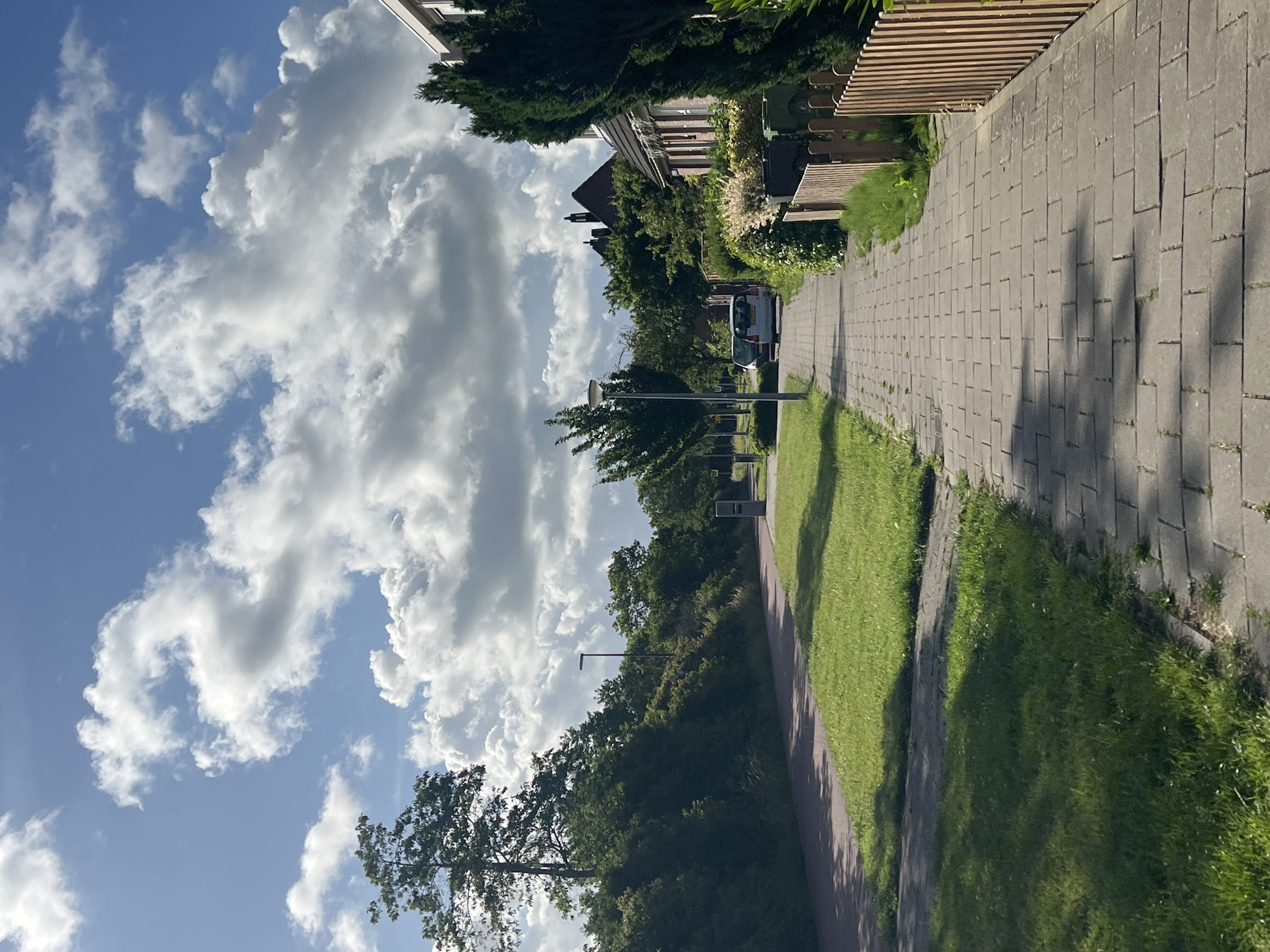 The image depicts a residential street in Alkmaar, Netherlands, on a sunny day with scattered clouds. A paved pedestrian walkway runs along the right side of the image, bordered by a wooden fence and lush greenery. Adjacent to the walkway is a red bicycle path, separating it from a strip of mown grass. Further to the left, dense vegetation and trees form a natural barrier. In the background, houses with red-tiled roofs are visible amongst the trees. Two cars are parked along the street. The sky is a vibrant blue, with large, puffy white clouds casting shadows on the ground. The overall scene conveys a sense of quiet suburban life.