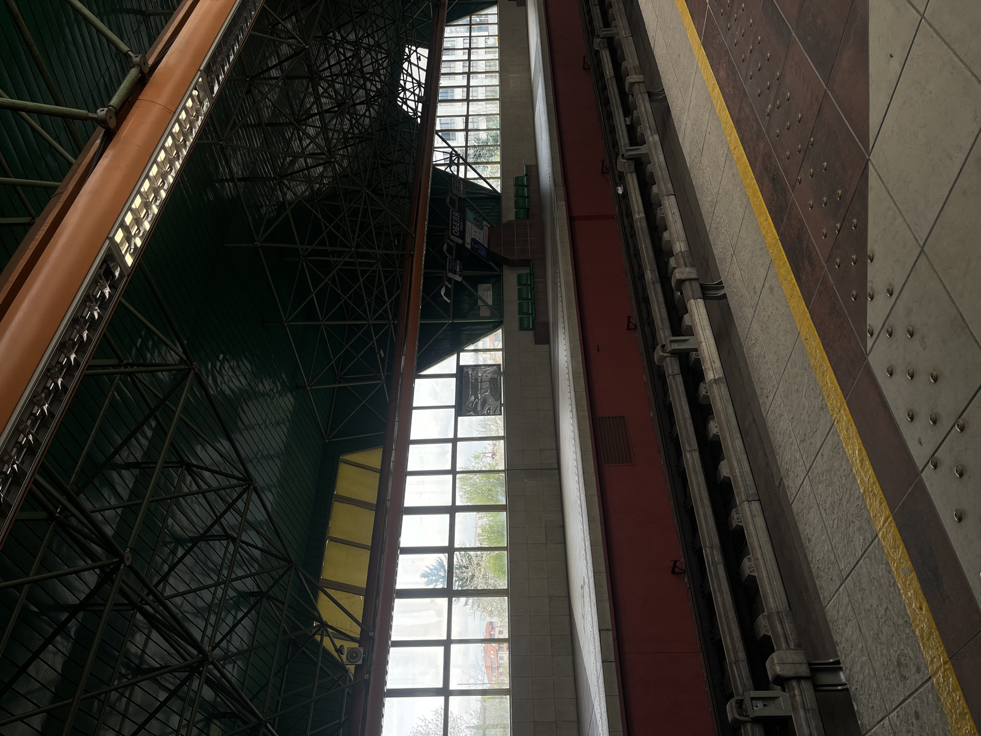 The image shows the Nadezhda metro station in Sofia, Bulgaria. The view is from a high vantage point, possibly an upper level or mezzanine, looking down on a portion of the platform. To the left is the station's roof structure, a grid of metal beams and a green ceiling. A long orange structure with lights runs along the roofline. Large windows are visible, offering a glimpse of the outside, including buildings and trees in bloom, suggesting springtime. In the center is a wall with green chairs and signs. To the right is the platform edge, marked with a yellow line, and a wall with visible hardware. The lighting appears to be a combination of natural light from the windows and artificial light from the overhead fixtures.