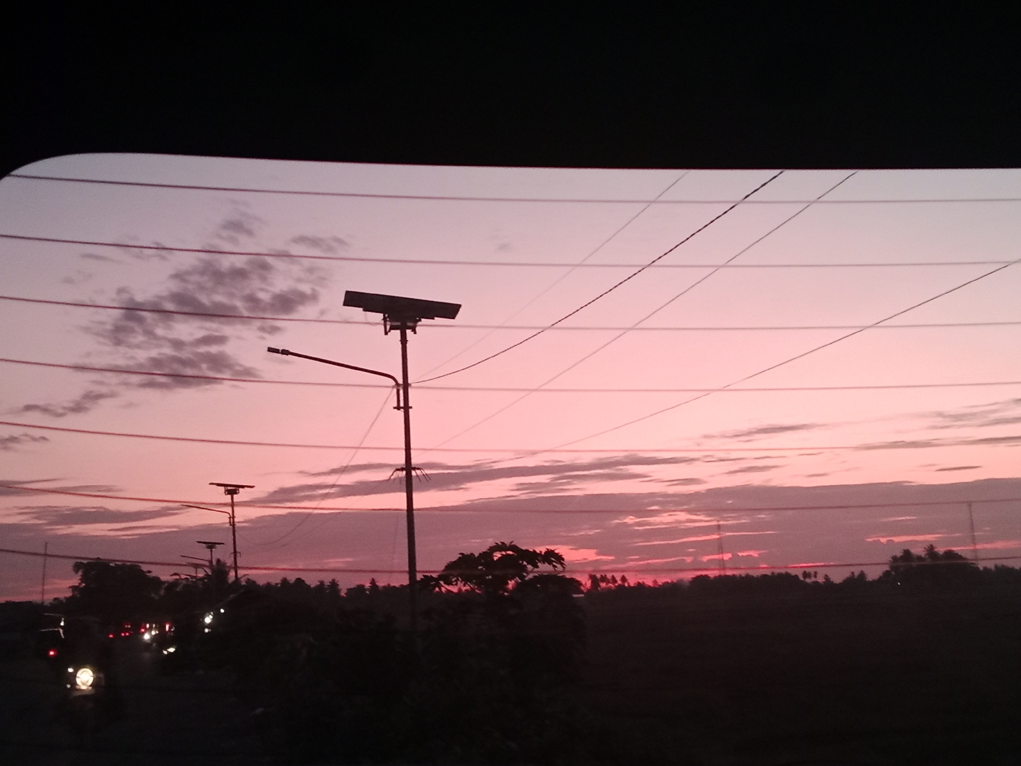 Captured through a rear window, the image showcases a dusky sky in Cotabato, Indonesia. The sky is painted with hues of pink and traces of cloud formations, and there are several power lines crisscrossing the view. Silhouetted against the horizon is a line of trees and foliage. In the foreground, there are two street lights, one of which features a rectangular solar panel. Further to the left, there appear to be car headlights, suggesting traffic on the road. The scene is viewed through a car window with horizontal defogger lines, and the vehicle frame partially obscures the upper portion of the view.
