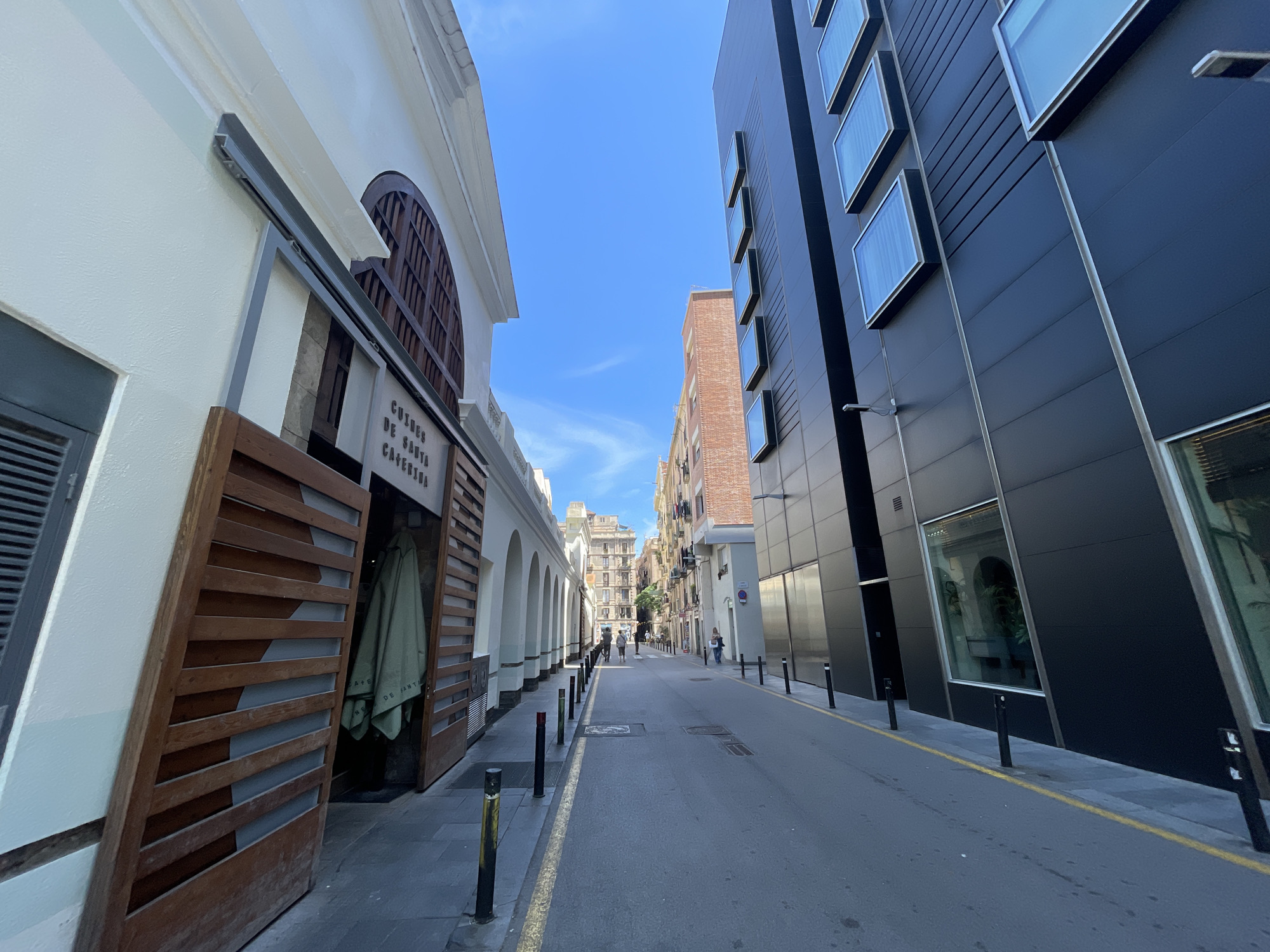 This detailed image captures a narrow, sunlit street in the city of Barcelona, Spain, specifically identifiable by the "CUINES DE SANTA CATERINA" sign on the left building, placing it adjacent to the Santa Caterina Market. The scene is presented from a low-angle, wide perspective, looking down the street on a clear, bright day.
On the left side of the street, a building with a light cream or white facade is prominent. It features an entrance with large, partially open wooden louvered doors, behind which a green garment or umbrella hangs. Above this entrance, the sign "CUINES DE SANTA CATERINA" is clearly legible in dark lettering. Further down this building, a series of light-colored arches is visible.
On the right side, a contemporary building rises steeply, clad in a dark grey or black facade with distinct horizontal paneling. Multiple rectangular windows, framed in a lighter color and appearing slightly recessed or projecting, are arranged vertically along its surface. A street light fixture extends from this building's facade. Reflections of the opposite building's arches are discernible in the lower windows.
The street itself is asphalt, bordered on both sides by a continuous yellow line. Black, cylindrical bollards are regularly spaced along both sides, separating the roadway from the pedestrian sidewalks. Several manhole covers are visible on the street surface.
In the middle and far distance, several indistinct individuals are seen walking along the street. The street recedes into a cluster of older, multi-story buildings constructed of brick and stone, some featuring balconies, under a clear blue sky with a few faint, wispy clouds.