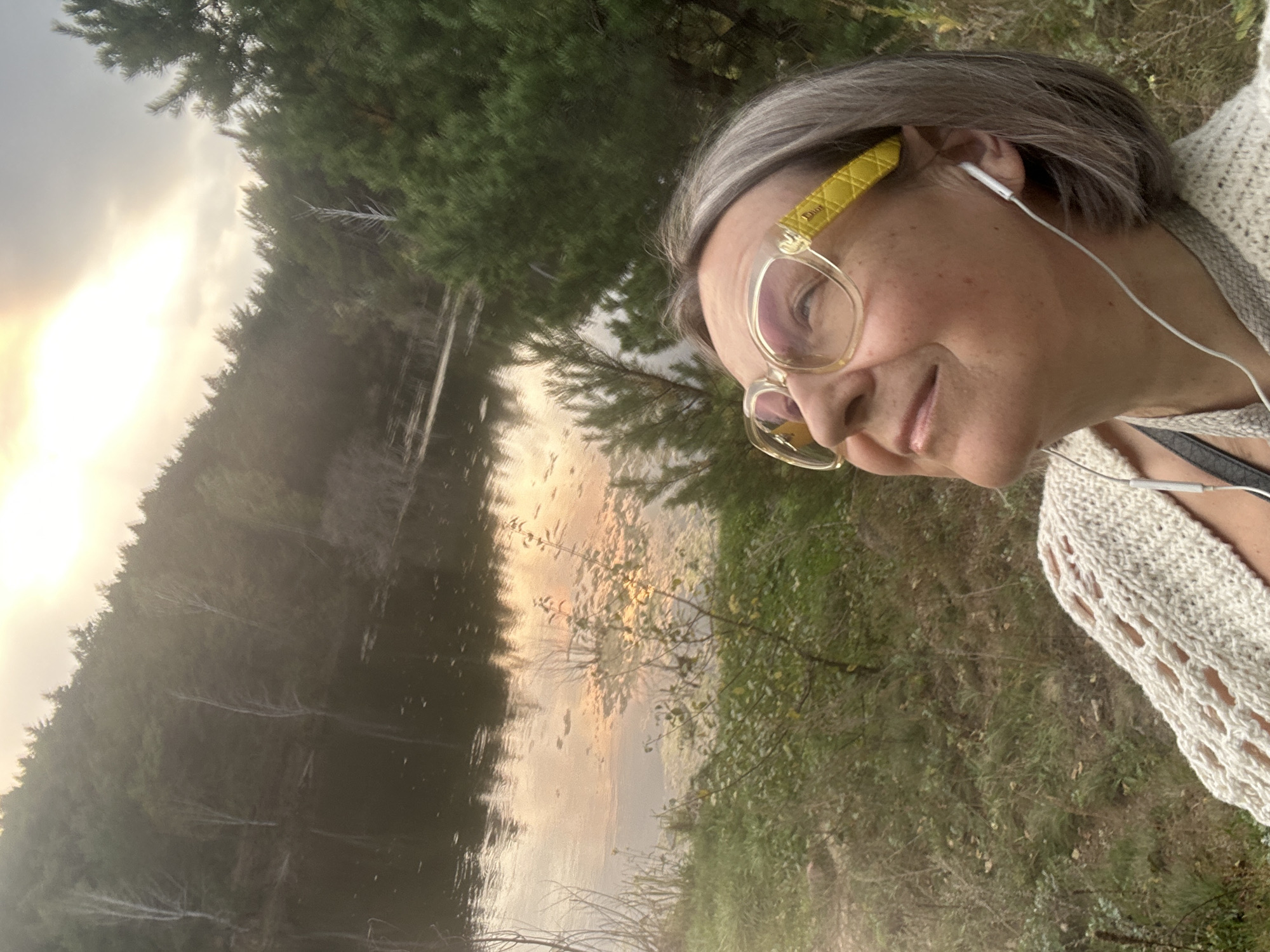 A woman with short gray hair and glasses is looking up at the sky. She is wearing a white knitted sweater and earbuds. She is standing in front of a lake and a forest. The sun is setting, casting a warm glow on the water and trees. The location is not specified.