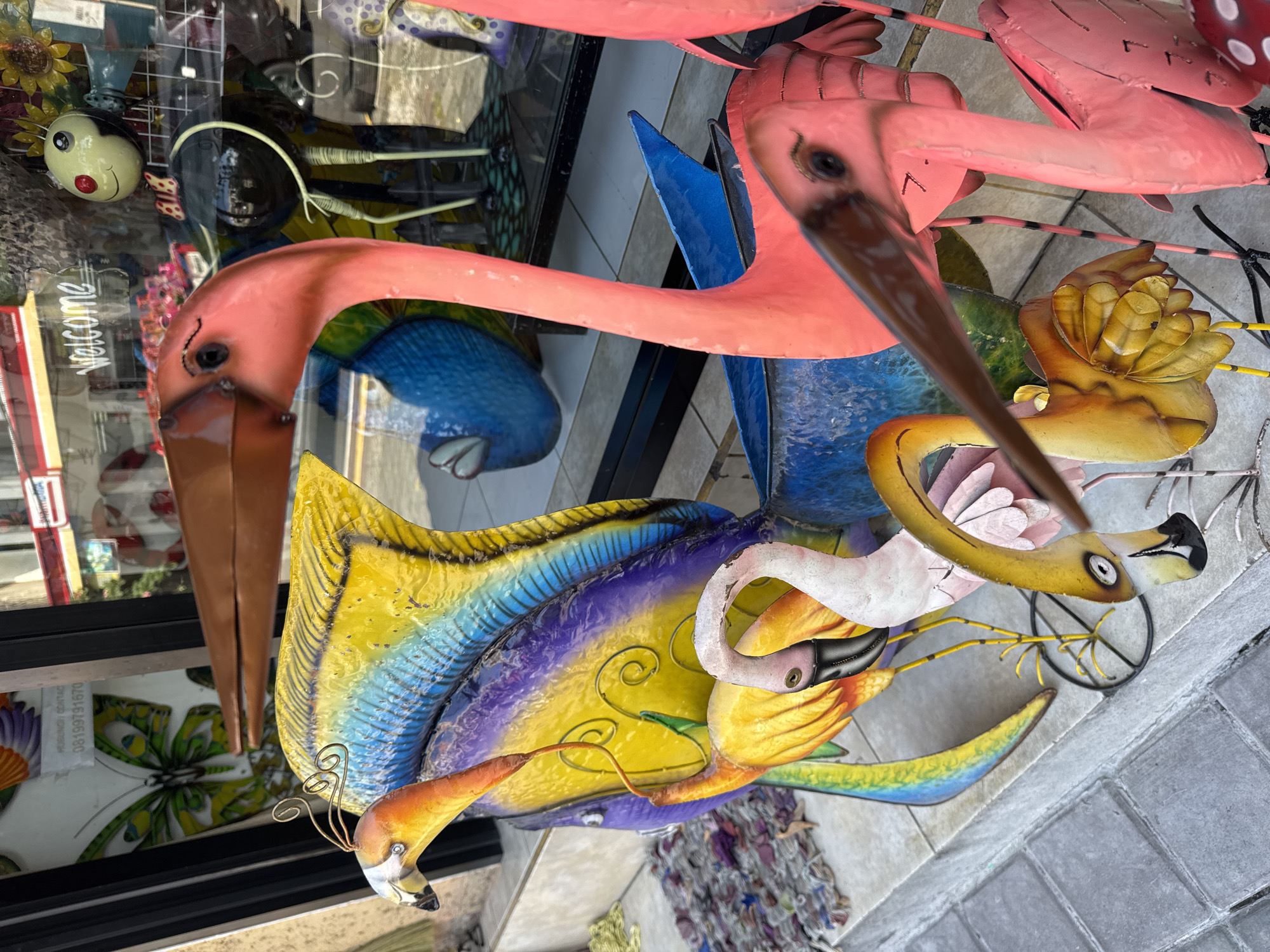 The image shows a collection of colorful metal flamingo sculptures displayed outside a shop. The sculptures vary in size and color, with some painted in vibrant shades of pink, yellow, blue, and purple. They are arranged on a paved sidewalk, with a shop window in the background displaying more items. A sign in the window reads "Welcome" and features a graphic of a butterfly with the text "MUBUNCH CONTACT 08199791670". The context suggests this is a street scene in Ubud, Indonesia, likely a souvenir shop or art gallery.