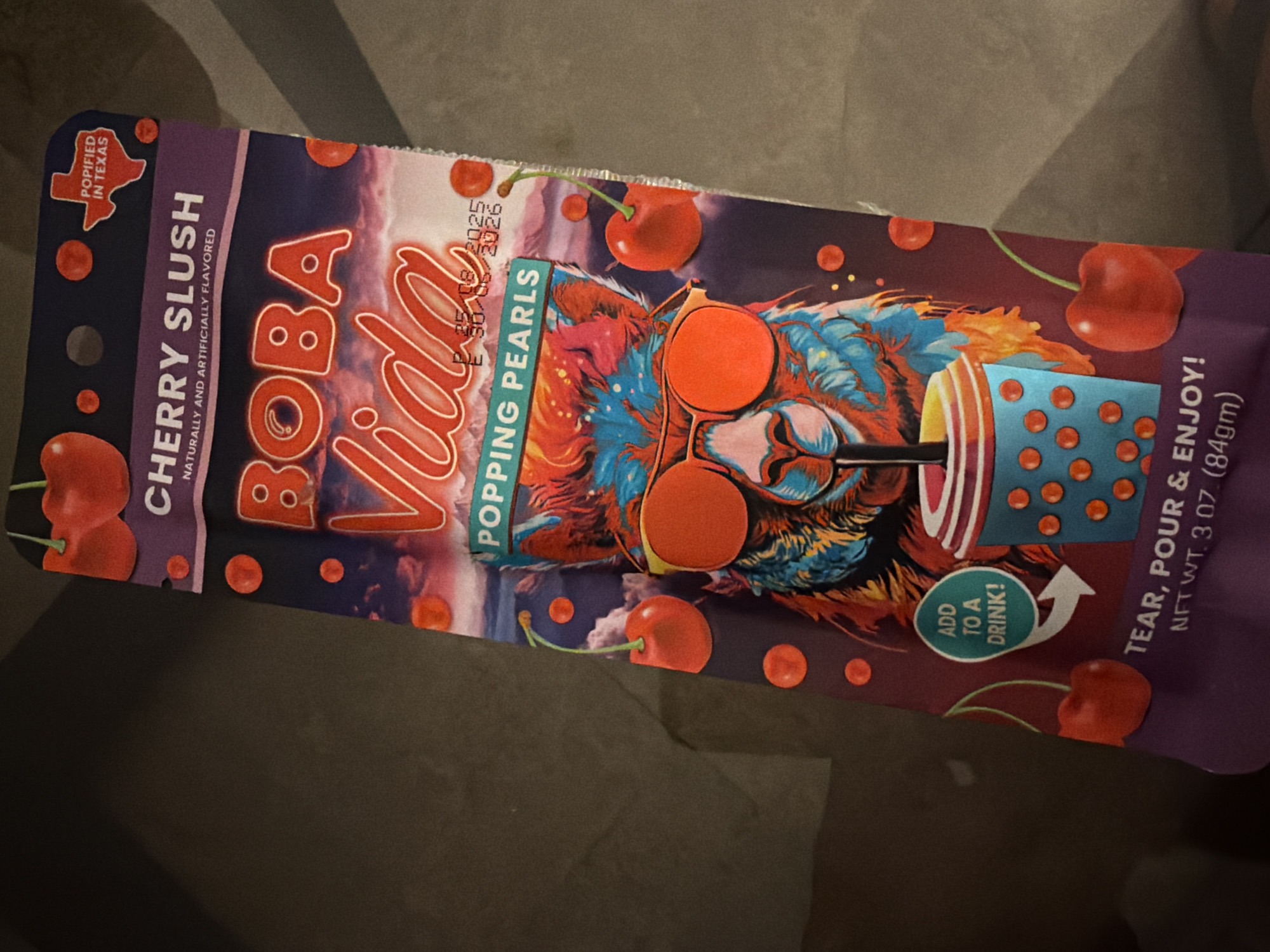 A product package for "Boba Vida Cherry Slush Popping Pearls" is displayed diagonally against a dark, textured surface. The packaging features a colorful illustration of a llama wearing sunglasses, holding a boba tea cup. The background of the package depicts a vibrant, cloudy sky with mountains. Text on the package indicates "CHERRY SLUSH NATURALLY AND ARTIFICIALLY FLAVORED," "POPPING PEARLS," and "TEAR, POUR & ENJOY! NET WT. 3 OZ (84gm)." There is also a small graphic of the state of Texas with the text "POPIFIED IN TEXAS." Expiration dates are printed in small text: "P 25/08/2025" and "E 30/08/2026". The image is taken from a medium close-up perspective, with the product occupying the majority of the frame.