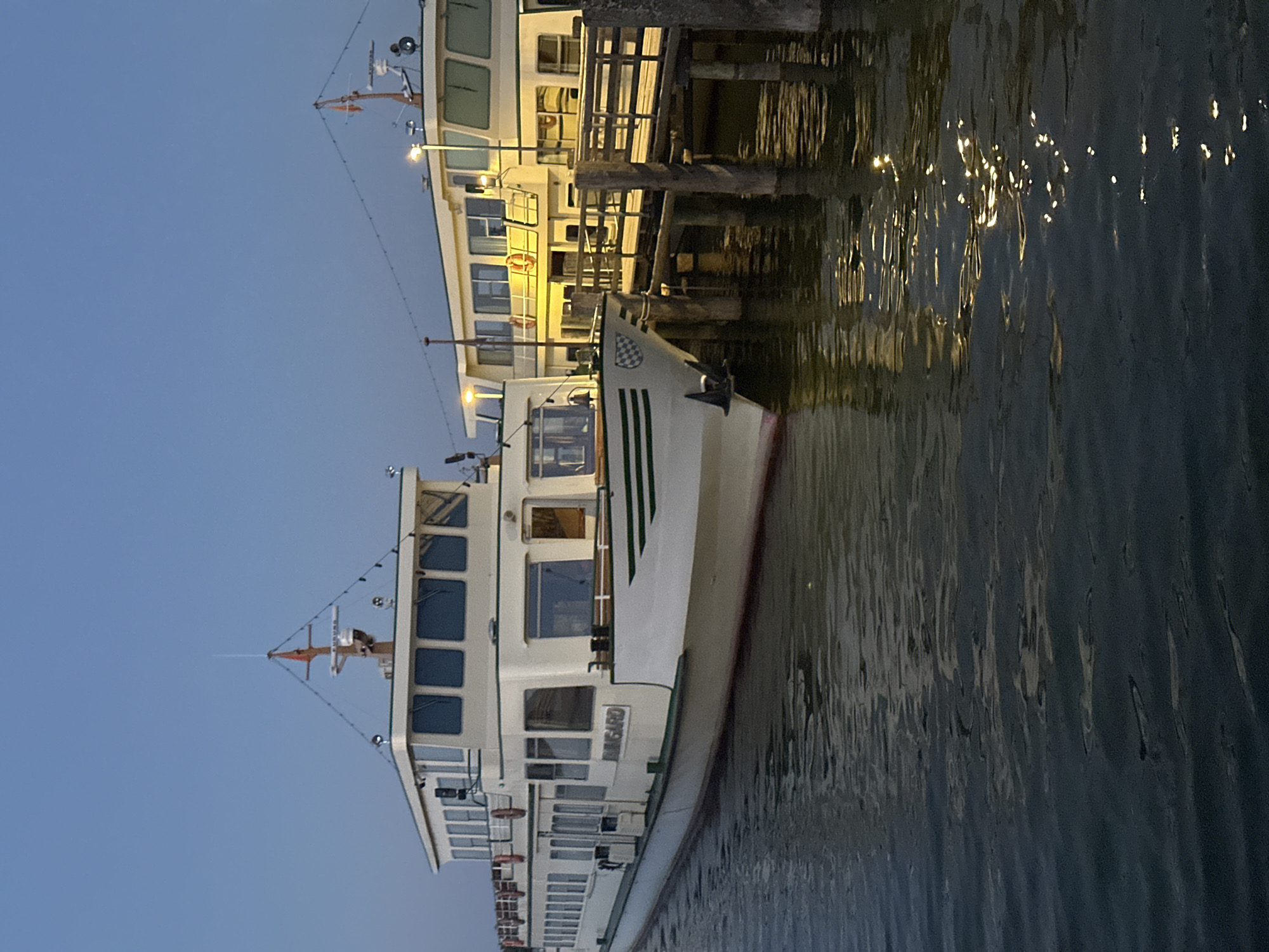 The image shows two passenger boats docked at a pier in Prien am Chiemsee, Germany. The boat in the foreground is white with green trim and has "RHEINGARD" written on its side. It has multiple windows and appears to be a ferry or tour boat. The second boat, partially visible behind the first, is also white and has illuminated windows, suggesting it is also a passenger vessel. The boats are moored to a wooden pier, which is illuminated by lights. The water is dark and reflects the lights from the boats and the pier. The sky is a clear, pale blue, indicating either dawn or dusk. The overall scene is serene and suggests a quiet moment before or after the boats have been in service.