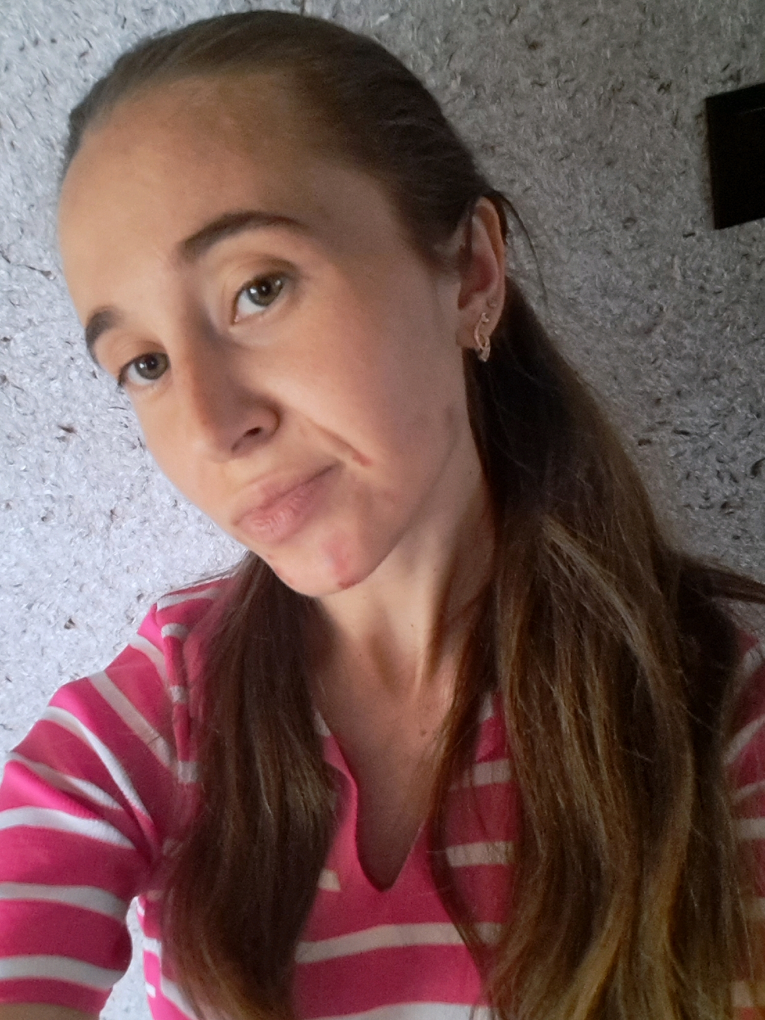 A woman is shown in a close-up, likely a selfie, wearing a pink and white striped shirt. Her hair is long and brown, and she is wearing earrings. There is a mark or scratch on her face near her mouth. The background appears to be a textured wall. The image does not provide any information about the location within the city of Vypasne, Ukraine, or any other environmental details.