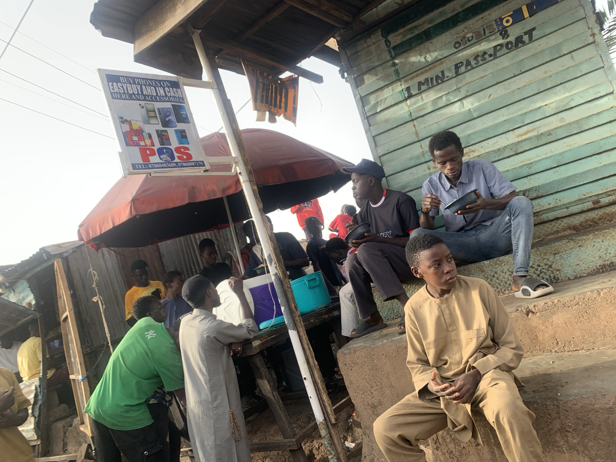 This media file depicts a lively outdoor scene in Jos, Nigeria, likely a street market or a commercial informal area during daylight hours, characterized by strong sunlight and clear weather. The setting features makeshift structures, including a corrugated metal building and a stall with a red umbrella for shade.
Numerous young men and boys are present. In the foreground, a boy in a light brown traditional kaftan sits on a concrete step, looking thoughtfully to his right. Behind him, two men are seated on a raised platform, possibly part of the corrugated metal structure, eating from bowls with spoons. One wears a light blue collared shirt and jeans, the other a dark t-shirt and cap. Other individuals are standing around a wooden stall, some observing, some possibly engaged in transactions or conversation. One man in a green t-shirt is seen from the back, and another in a light grey traditional outfit stands near a white cooler.
Notable objects include commercial signs attached to the structures. One sign, white with blue text, advertises "BUY PHONES ON EASYBUY AND IN CASH HERE AND ACCESSORIES." It also features "POS" and two telephone numbers: "07060403609" and "07068087179." Another sign, hand-painted on the corrugated metal wall, reads "1 MIN. PASS-PORT," indicating a service for quick passport photographs or related documentation. A light blue plastic container and a white cooler are visible on a wooden counter.
The overall atmosphere suggests a casual, bustling environment where daily life and commerce intertwine, characteristic of informal markets in urban settings. The structures are practical and utilitarian, reflecting common adaptations in such locales.