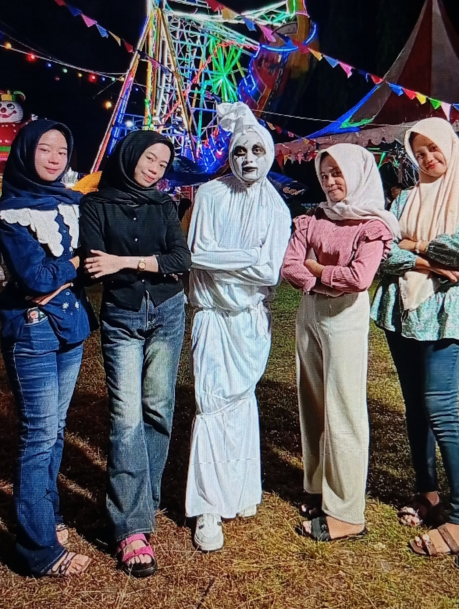 Five individuals are posing for a photograph in an outdoor setting at night. The background features a brightly lit amusement park ride with colorful lights and triangular flags. The ground is covered in dry grass.
From left to right, the individuals are:
1. A woman wearing a dark blue hijab and a denim jacket with white ruffled details. She is wearing blue jeans and sandals.
2. A woman wearing a black hijab and a black button-up shirt. She is wearing blue jeans and pink sandals.
3. A person in a white costume that covers their entire body, resembling a ghost. The face is painted white with dark eye makeup and a black mouth. They are wearing white shoes.
4. A woman wearing a light pink hijab and a pink long-sleeved shirt with ruffled shoulders. She is wearing light-colored, wide-legged pants and black sandals.
5. A woman wearing a light beige hijab and a patterned long-sleeved shirt with a light green and blue print. She is wearing dark blue jeans and brown sandals.
The individuals appear to be at an event or festival. The presence of the amusement park ride and the festive decorations suggest a public gathering. The location is identified as Lhokseumawe, Indonesia, implying the photograph was taken within or near the city.