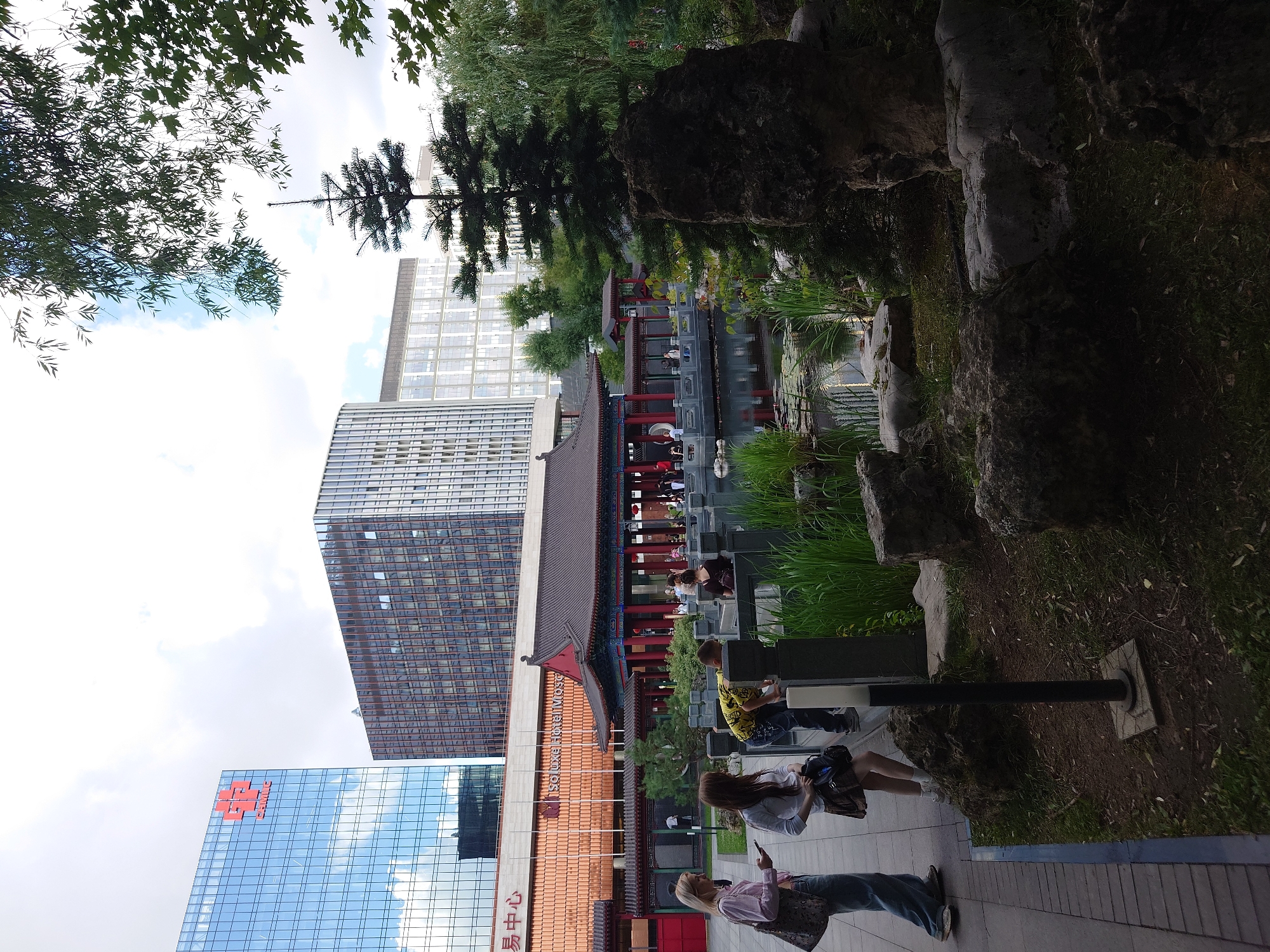 The image depicts the central courtyard of the Moscow Chinese Garden of Friendship, located within the Aptekarsky Ogorod botanical garden of Moscow State University. Two young women and a young boy are interacting near a pond. One woman, on the left, is wearing a pink button-up shirt and blue jeans, holding a smartphone. The other woman, next to her, wears a white shirt and a brown skirt, carrying a dark bag. The boy, wearing a yellow shirt with a black print, leans over a railing, looking into the pond. In the background, a traditional Chinese pavilion with red columns and a dark roof is visible. Behind the pavilion, modern glass-fronted skyscrapers rise into a cloudy sky. The foreground features a naturalistic garden with rocks, grass, and tall reeds.