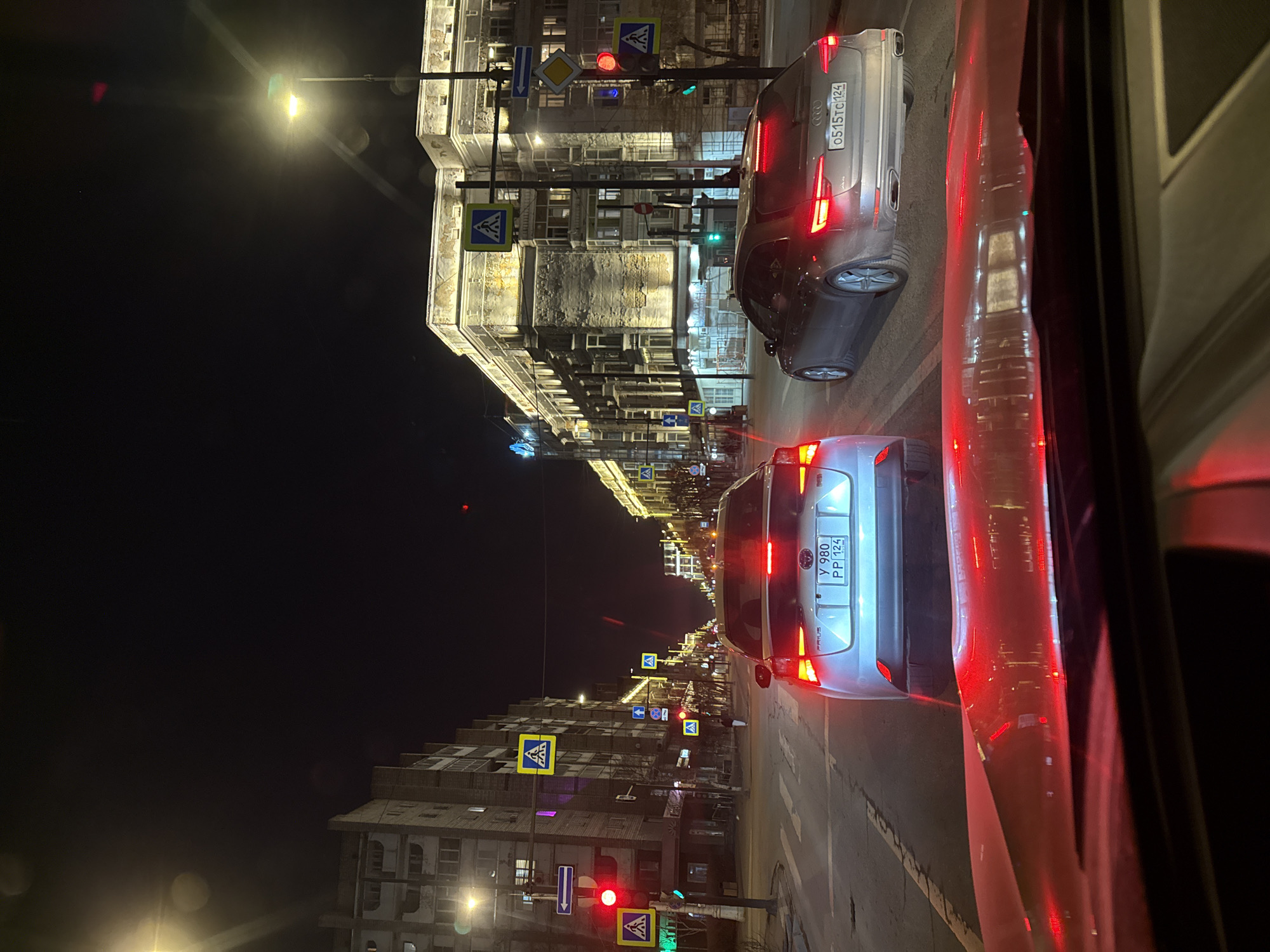 This is a nighttime photograph depicting a busy city street from the perspective of a vehicle, looking out the side window. The scene is dominated by artificial light sources, including streetlights, car lights, and illuminated building windows.
In the midground, two cars are prominently featured with their red brake lights illuminated, indicating they are stopped or moving slowly in traffic. The vehicle closest to the viewer is a white car, possibly a sedan, with a Russian license plate "Y 980 PP 124". Ahead and slightly to its left is a silver Audi, displaying the license plate "O515TC124" and an Audi logo on its trunk. The "124" region code on both plates confirms the location as Krasnoyarsk Krai, Russia. Reflections, predominantly red, from the interior of the vehicle taking the photo are visible on the right side of the image.
The street is lined with multi-story buildings, some appearing older with intricate facades, while others are more modern. Many windows are lit, adding to the urban nighttime atmosphere. Numerous street signs are visible, including multiple blue square signs with white pedestrian crossing symbols, a yellow diamond-shaped 'priority road' sign, and a blue rectangular 'straight ahead' sign. Traffic lights showing both red and green signals are also visible further down the street. Overhead, bright streetlights cast a yellow-white glow, with a distinct lens flare in the upper left of the frame. The overall scene captures the vibrant activity of an urban center after dark.