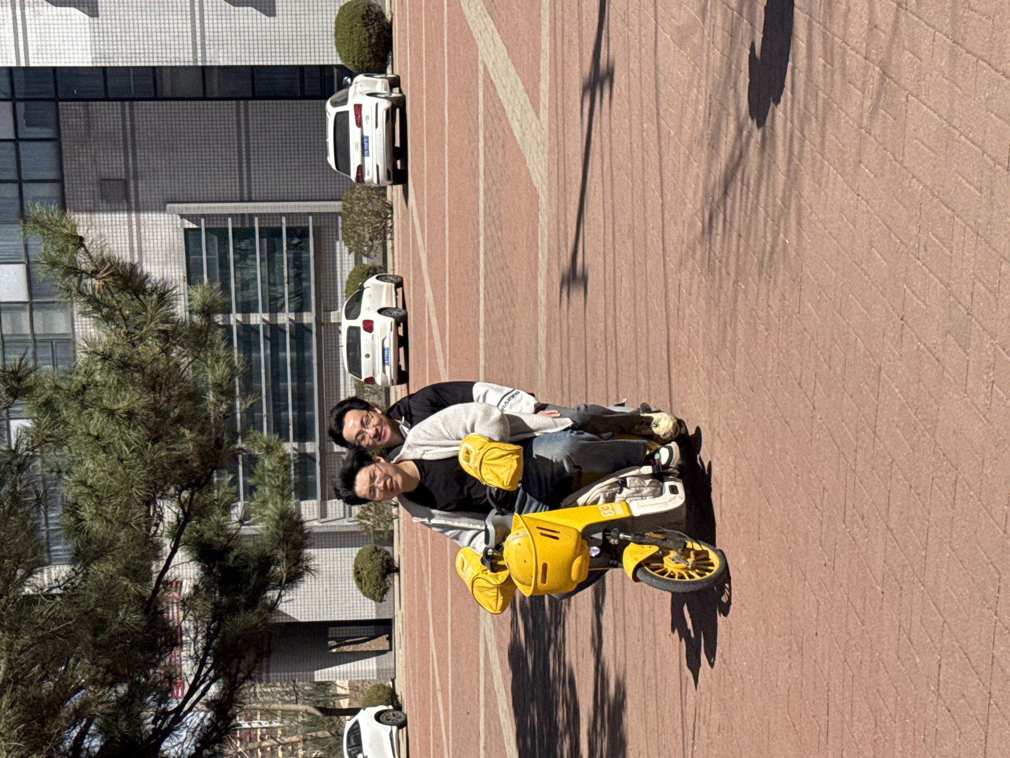 Two men ride a yellow scooter on a brick-paved surface in Zhongtun, China. A building with large windows and several white cars are in the background. A tree is on the left side of the image. A shadow of a pole is cast on the brick surface.