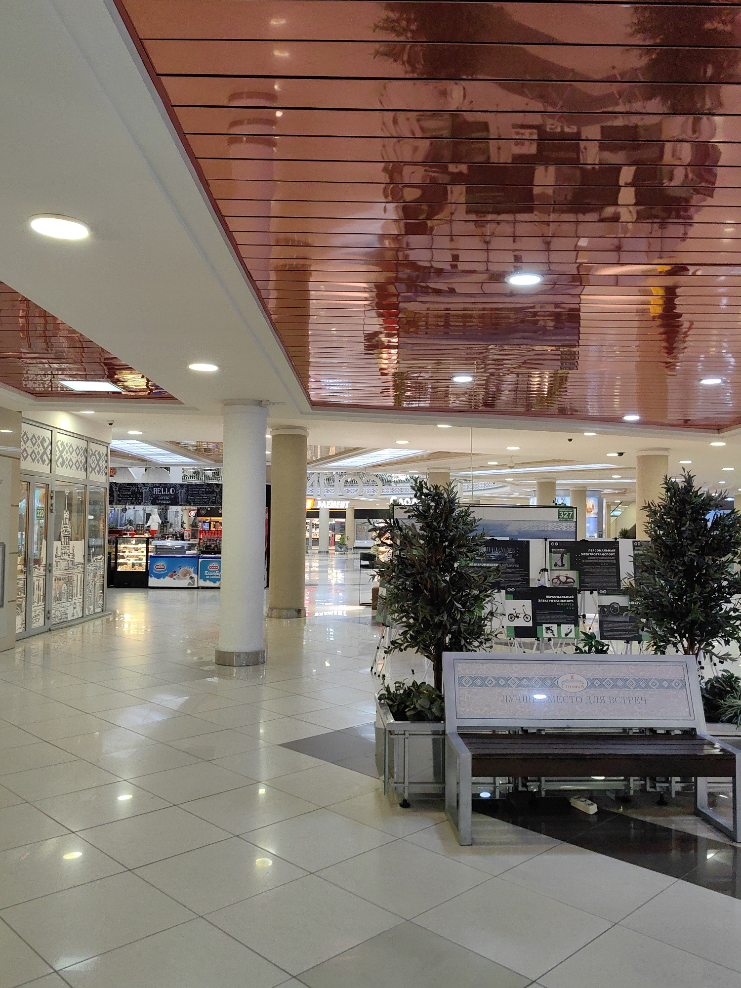 The image shows an indoor scene, likely a shopping mall or a large commercial space in Minsk, Belarus. The ceiling is a distinctive copper-toned, reflective material with a slatted design, creating reflections of the environment. The floor is tiled with large, light-colored square tiles, which are also reflective.
In the foreground, a bench with a dark wooden seat and a metal frame is visible. The back of the bench features a decorative panel with a light blue and white patterned design and text that appears to be in Cyrillic. Flanking the bench are two artificial potted trees.
The middle ground reveals a section of the mall with shops and kiosks. One shop has a glass facade with decorative elements and the number "320" visible. Adjacent to it, a food or beverage kiosk displays pastries in a refrigerated case and has signage that includes "HELLO COFFEE." Further in the background, more shops and architectural elements of the mall are visible, including large columns and illuminated signage. Two other columns, one white and one beige, are prominent in the mid-right of the image.
The background shows more of the mall's interior, with a high, illuminated ceiling and various retail spaces. The overall impression is of a clean, modern, and well-lit commercial environment. There are no people clearly visible in the image. The reflections on the ceiling and floor suggest a brightly lit space, possibly with overhead lighting and natural light filtering in.