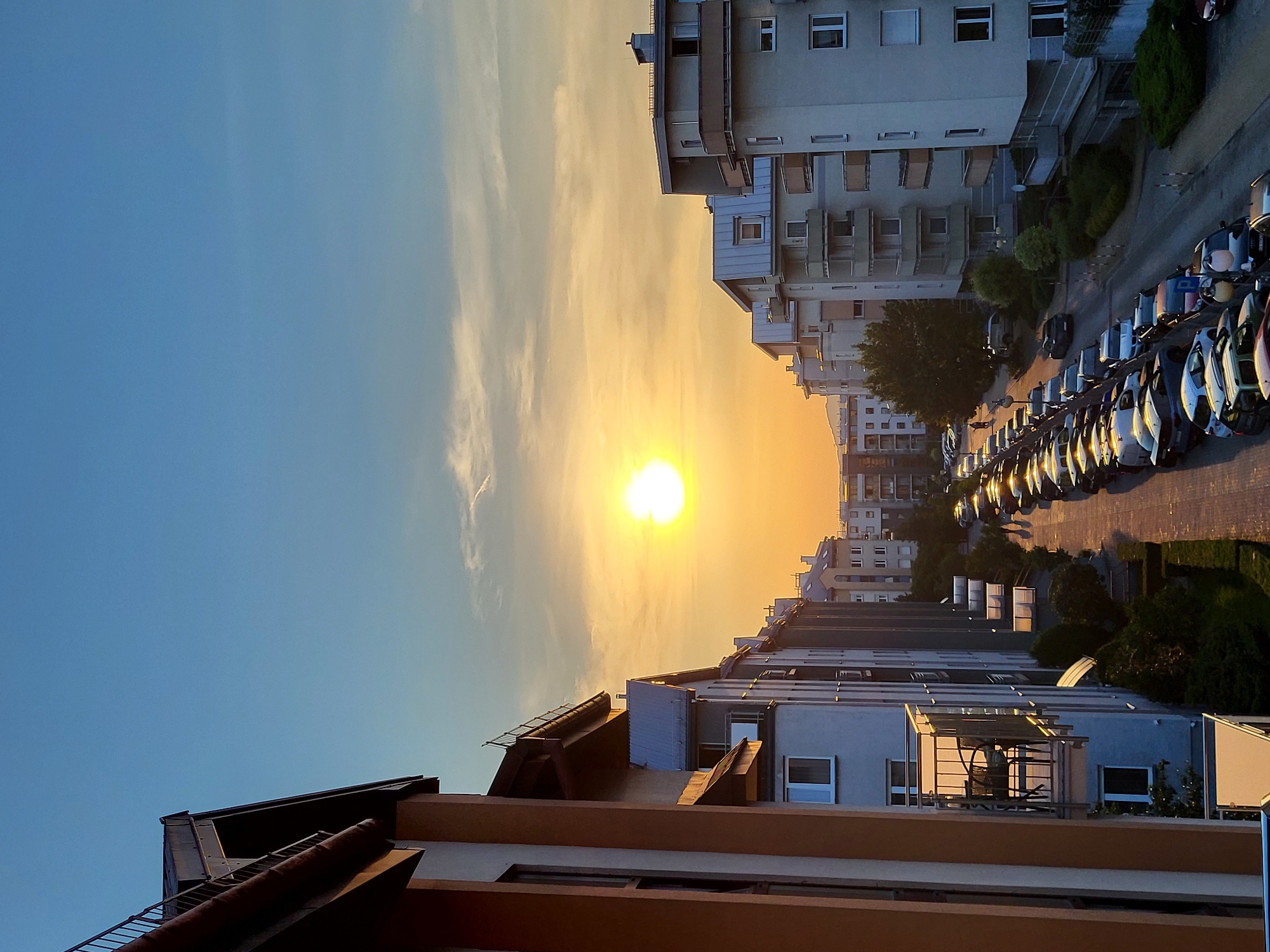 The image shows a street lined with apartment buildings at sunset. The sun is low in the sky, casting a warm orange glow. A row of cars is parked along the side of the street. The buildings appear to be modern, with balconies and large windows. The sky is a gradient of blue and orange, with a few wispy clouds. There are a few scattered trees and green bushes along the sidewalk. The perspective is from a higher vantage point, looking down the street.