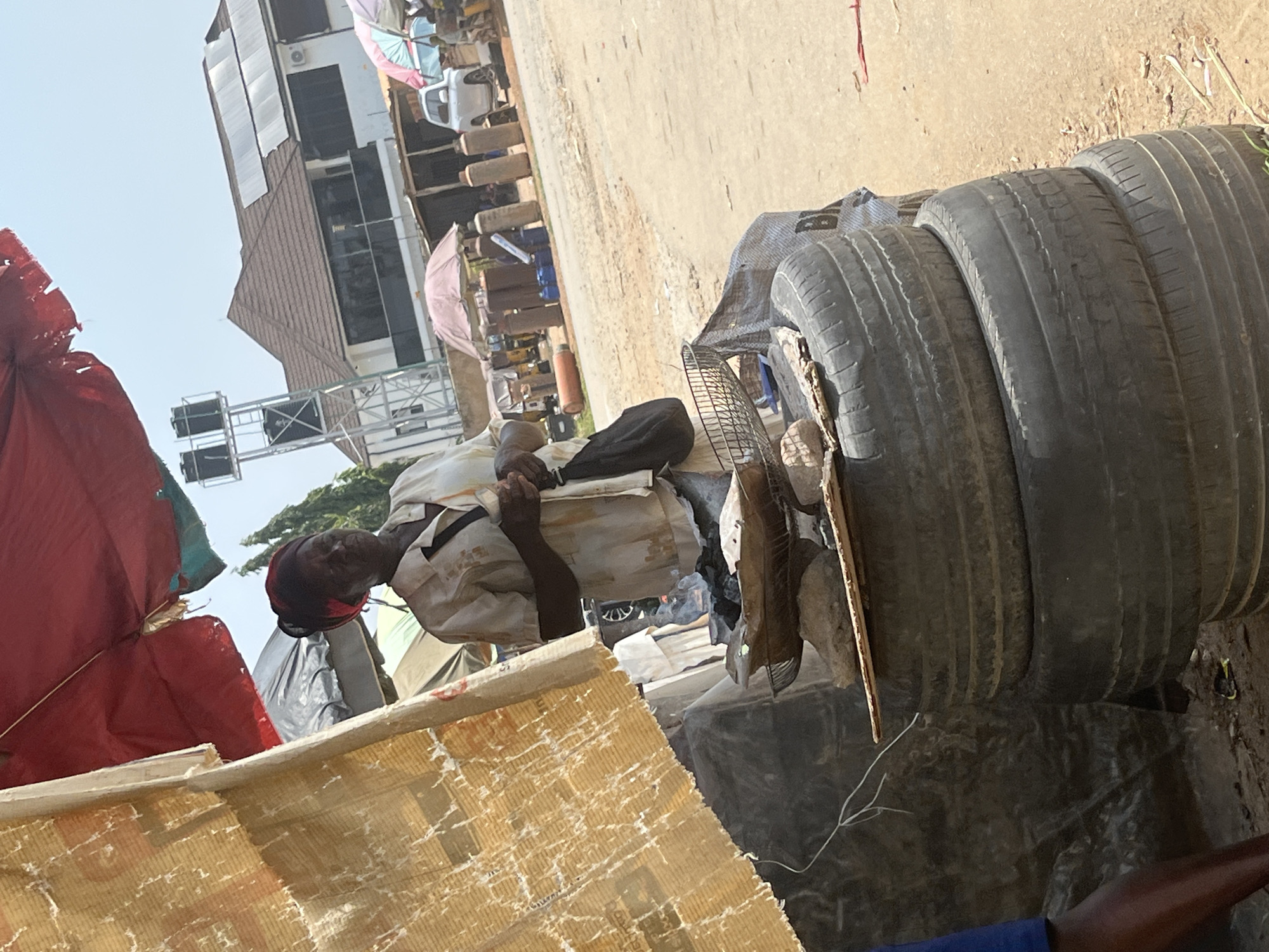 The image depicts an outdoor scene in Abuja, Nigeria, likely during daylight hours given the bright illumination. The focal point is an elderly individual, presumably male, with dark skin and wearing a light-colored shirt and a dark head covering, engaged in reading a document. The person is seated amidst a collection of stacked tires, some of which are worn and weathered. A black bag is placed near the individual, and a wire fan is positioned in front of the tires.
The background reveals a somewhat bustling environment with elements suggesting a market or roadside stall. Various structures, including a multi-story building with a distinctive slanted roof and windows, are visible. Other objects, such as market stalls with colorful umbrellas (one pink and one lighter colored umbrella, and what appears to be a red tarpaulin), and what might be gas cylinders or tanks, are also present. The ground is a dusty, sandy surface.
The scene appears to be captured in an informal setting, possibly a place where old tires are recycled or sold. The overall impression is one of everyday life in an urban Nigerian environment. No visible text is discernible in the image.
