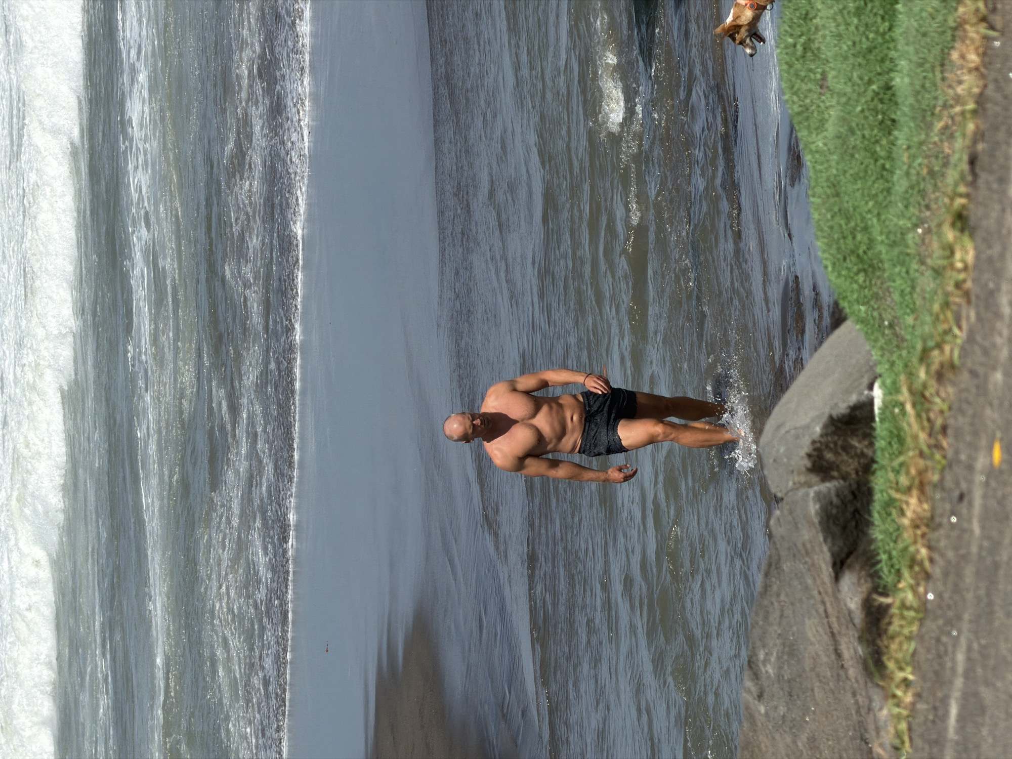 A bald, muscular man in black swim trunks walks out of the ocean at a beach in Mengwi, Indonesia. He is walking towards the viewer with his head slightly tilted. Behind him, waves crash on the shore. In the lower right corner, a small dog with a red collar looks out towards the water. The environment is a sandy beach with rocks and grass in the foreground. The sky is a hazy gray.