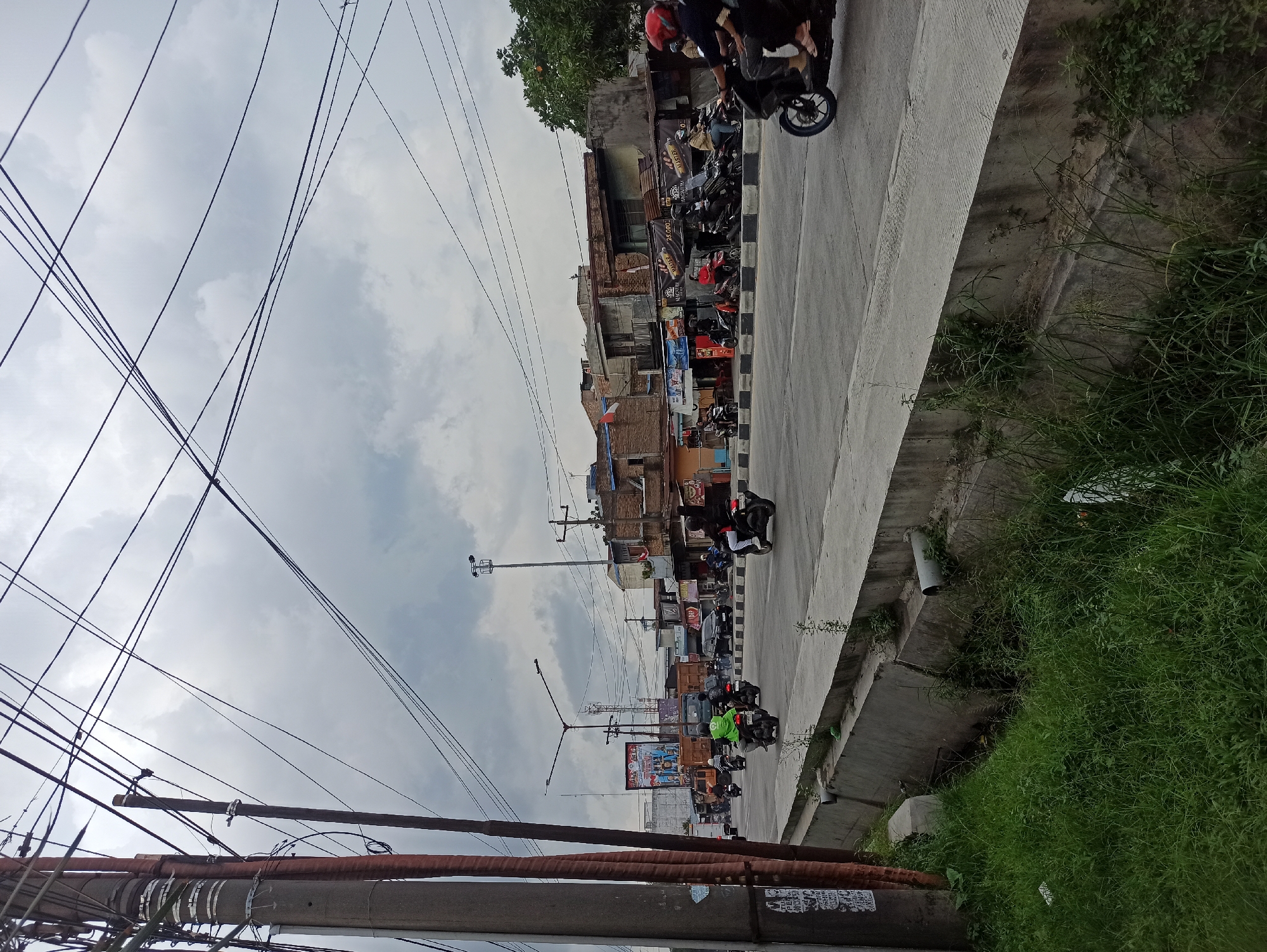 The image depicts a street scene in Medan, Indonesia, under an overcast sky. The foreground is dominated by a concrete drainage ditch filled with overgrown grass and vegetation, with a concrete barrier separating it from the road. A utility pole with numerous wires runs vertically along the left edge of the frame. The road itself is paved and carries moderate traffic, primarily consisting of motorcycles. Several individuals are seen riding motorcycles, some with passengers. On the right side of the road, there is a row of low-rise buildings, appearing to be shops or residences, with some storefronts visible. In the background, there are more buildings and a large billboard. The overall atmosphere is that of a busy urban environment.