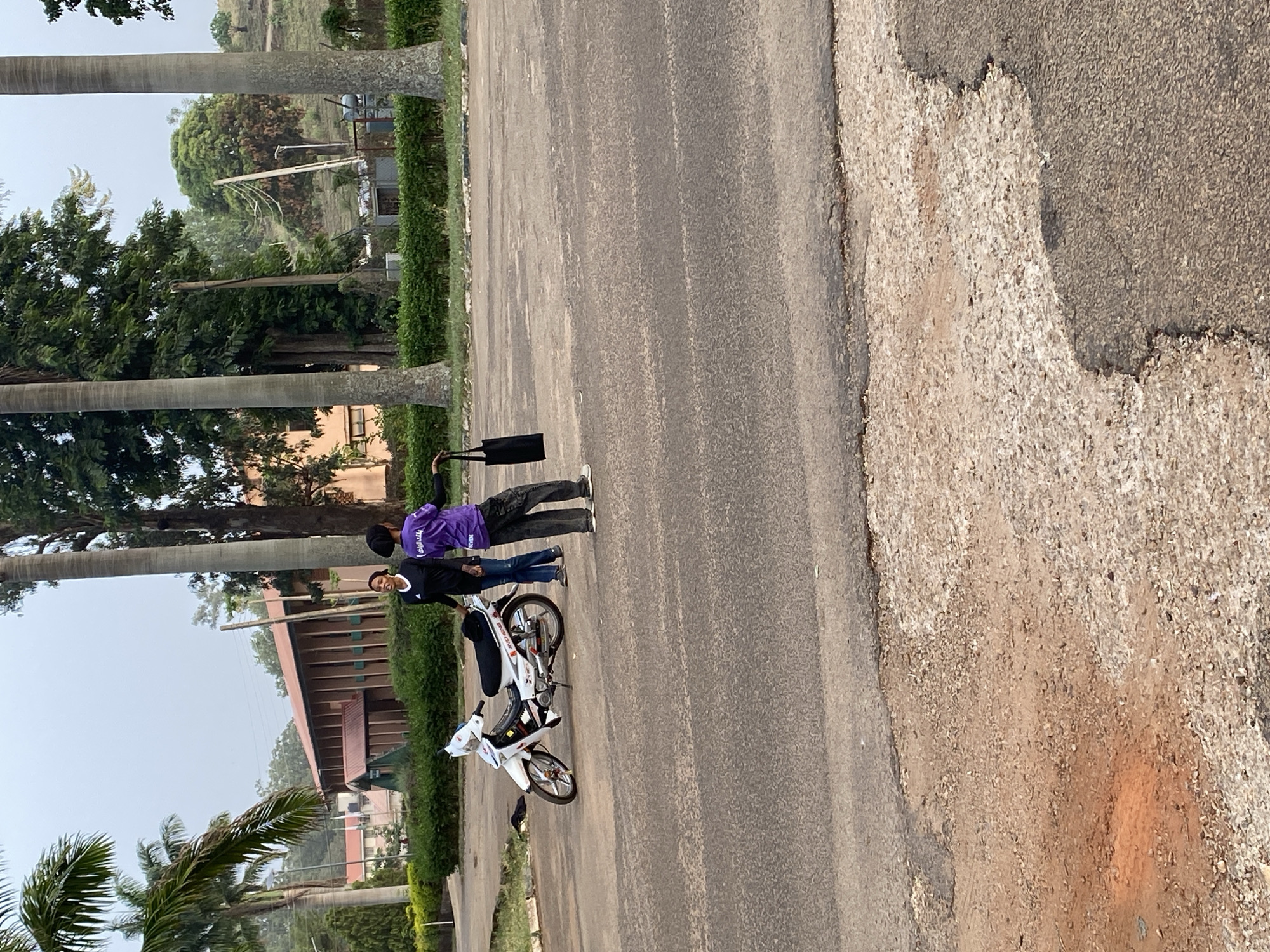 The image, taken in Vom, Nigeria, captures an outdoor scene featuring two individuals and a white motorcycle on an asphalt road, bordered by green foliage and distant buildings. The photo is oriented vertically, suggesting a portrait-mode shot rotated sideways.
The main subjects are two people standing on the paved road. On the left, an individual wears a purple long-sleeved top with white text (partially visible, possibly "MALARIA" and other words) and dark trousers, holding a black shoulder bag. To their right, another individual wears a dark top, possibly black with white trim, and blue jeans. This person is standing next to a white step-through motorcycle, which has "JINGCHENG" written on its side panel. This individual is smiling towards the camera.
The setting appears to be a campus, park, or institutional area. The road is asphalt, with a distinct lighter-colored, rougher gravel and dirt shoulder to the right. Tall palm trees line the left side of the road, behind a well-maintained green hedge. Further back, numerous other green trees and bushes are visible, along with multi-story buildings featuring reddish-brown roofs and walls, partially obscured by the vegetation.
The sky is light and appears overcast or hazy, indicating daytime without direct sunlight, possibly early morning or late afternoon. The overall atmosphere is relaxed, with the individuals seemingly engaged in conversation or posing.