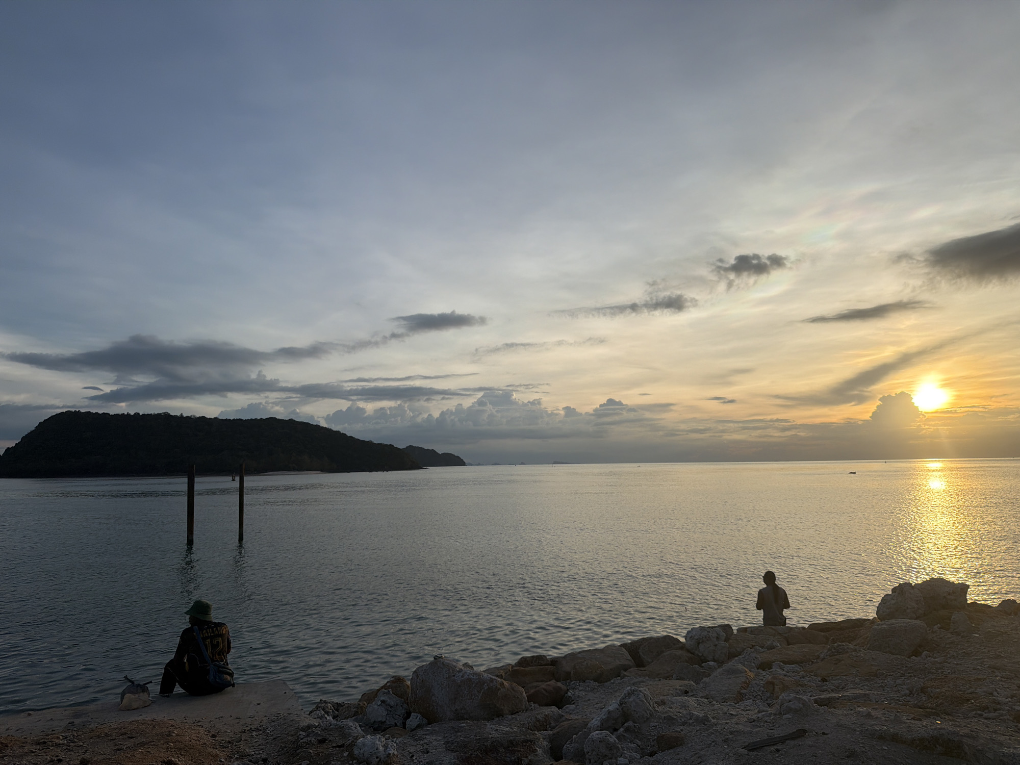 The image depicts a tranquil scene of a beach at sunset in Koh Phangan, Thailand. Two individuals are present, each seemingly enjoying the serene atmosphere. One person, wearing a hat, sits on the shore, facing the water and a distant island. Another person is positioned on a rocky outcrop, also facing the vast expanse of the sea. The sun is low on the horizon, casting a warm, golden glow across the water and the sky, suggesting the time of day is late afternoon or early evening. The sky is a blend of soft blues and oranges, with scattered clouds adding depth to the vista. The water is calm, reflecting the colors of the sky and the setting sun. A silhouetted island can be seen in the distance, adding a sense of isolation and natural beauty to the scene. The foreground consists of rocks and sand, typical of a coastal environment. The overall mood of the image is peaceful and contemplative, capturing the beauty of a sunset by the sea in a tropical location. There is text visible on the back of the shirt of the person sitting on the shore, which appears to be a number "12" and some illegible lettering.