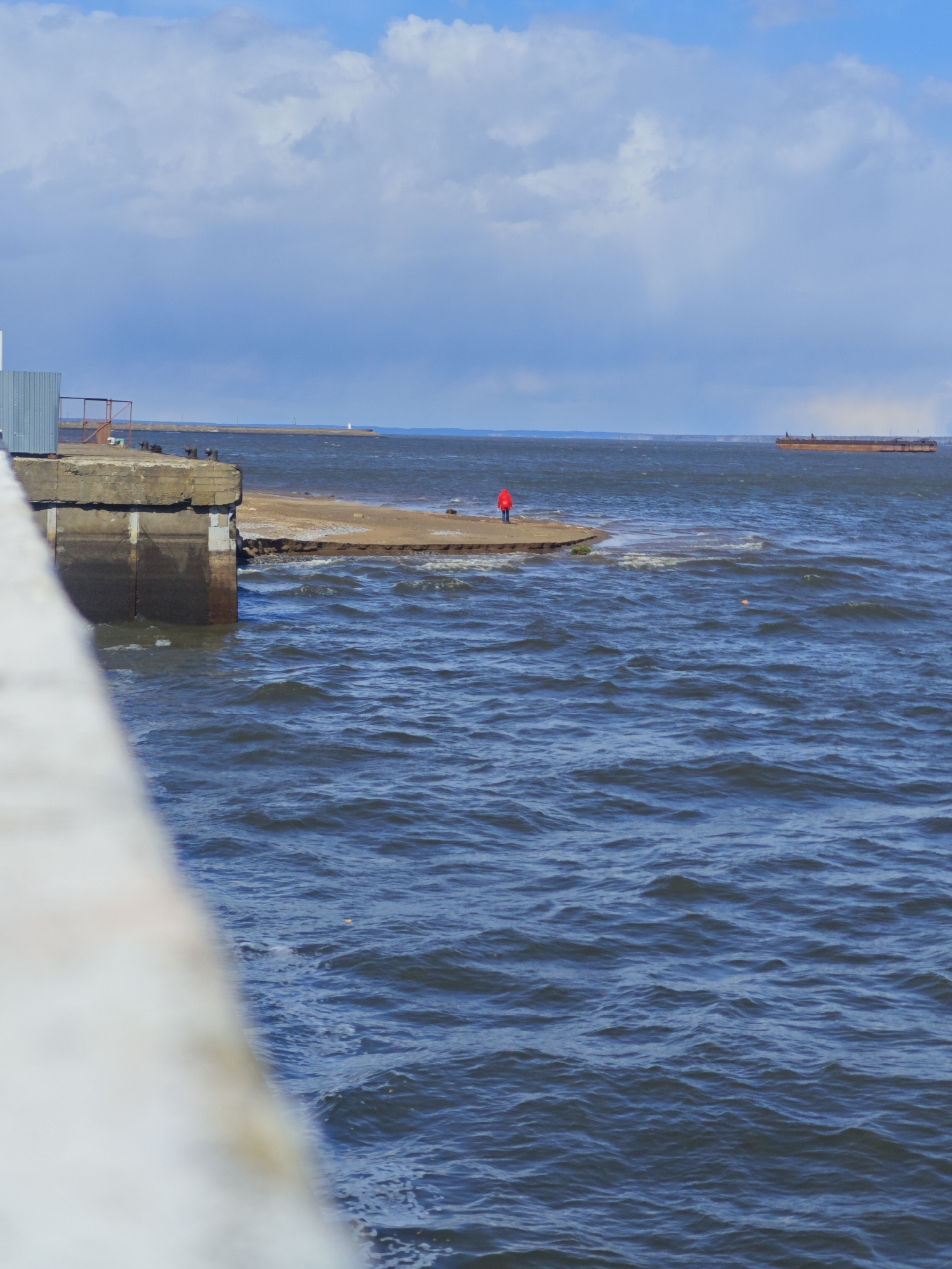 The image captures a vast expanse of choppy, dark blue water under a bright sky with scattered white clouds, likely during daytime in Chaikovsky, Russia. In the midground, a lone individual wearing a striking red jacket stands on a narrow, sandy strip of land that extends into the water. The person is facing away from the viewer, looking out towards the distant horizon.
To the left, a robust concrete pier or embankment occupies the foreground, featuring a light blue corrugated metal structure and several mooring bollards. The water laps and breaks against the base of this concrete structure and the adjacent sandy patch. The photograph appears to be taken from an elevated position, possibly from an overhead bridge or a higher section of a pier, with a blurred, light-colored structure in the immediate left foreground.
In the distance, across the expansive water, a long, flat-bottomed cargo barge is visible, either in motion or anchored. Further along the distant shoreline, a small white lighthouse can be seen on what appears to be a breakwater or a far-off landmass. The agitated surface of the water, with numerous ripples and waves, suggests windy conditions. No visible text is present in the image.