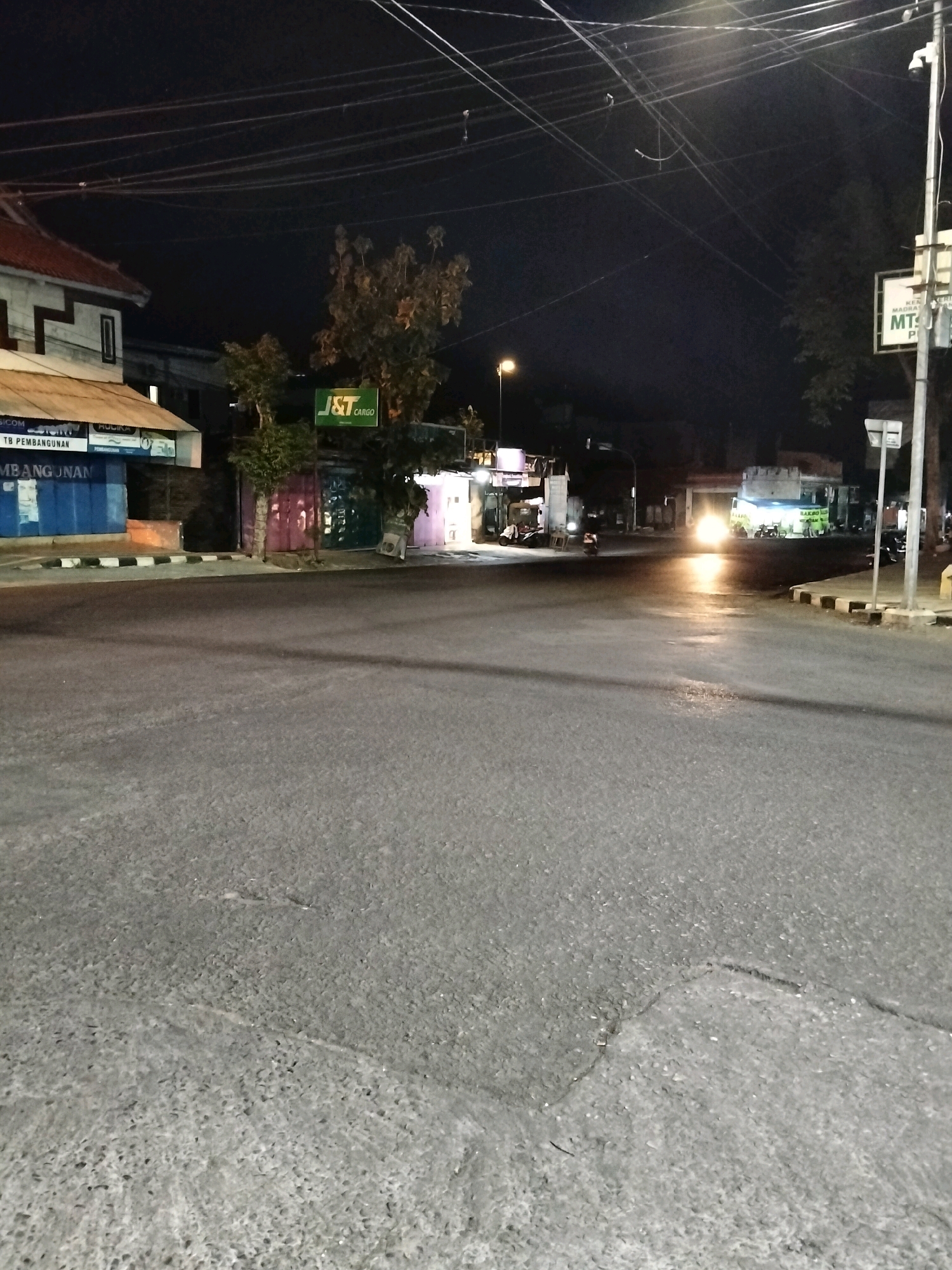 The image depicts a street intersection at night in Purworejo, Indonesia. The scene is illuminated by streetlights and the headlights of a vehicle. On the left side of the street, there are several buildings, including a structure with a white facade and a red roof, and another with blue and white siding. A green sign with "J&T CARGO" is visible on a building in the middle ground. Further down the street, there are more buildings and shops, with lights emanating from them. A streetlamp is also visible, casting a warm glow. The road surface appears wet, suggesting recent rain. A few motorcycles are parked near the shops. In the distance, a vehicle's headlights are a prominent source of light. The overall atmosphere is that of a quiet, urban night scene.