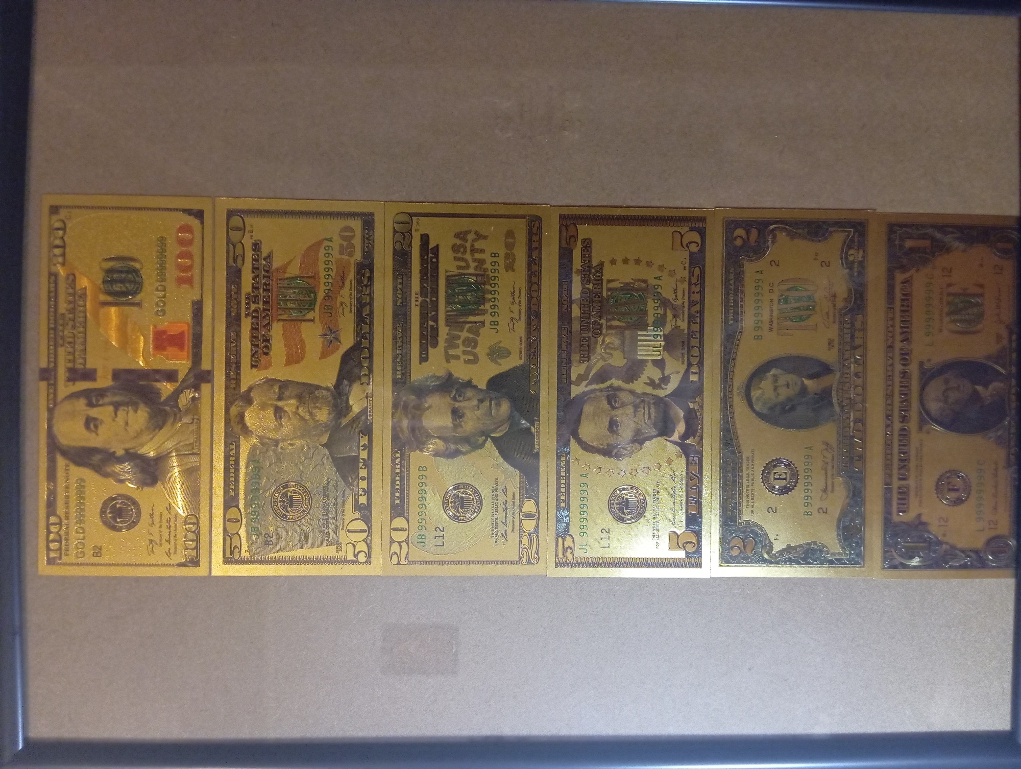 This media file displays a collection of gold-plated novelty dollar bills framed horizontally. The bills appear to be replicas of US currency and include denominations of $100, $50, $20, $5, $2, and $1. Each bill features a portrait of a historical figure, with Benjamin Franklin on the $100 bill, Ulysses S. Grant on the $50 bill, Andrew Jackson on the $20 bill, Abraham Lincoln on the $5 bill, Thomas Jefferson on the $2 bill, and George Washington on the $1 bill. Many of the bills have the serial number "99999999" with a letter prefix and suffix. The setting appears to be indoors, with the framed bills resting against a plain, light-colored surface. There are no indications of time of day, weather, or strong emotions. The visible text consists of the denominations, "UNITED STATES OF AMERICA," "FEDERAL RESERVE NOTE," and the serial numbers. The overall impression is that of a decorative display of novelty currency.