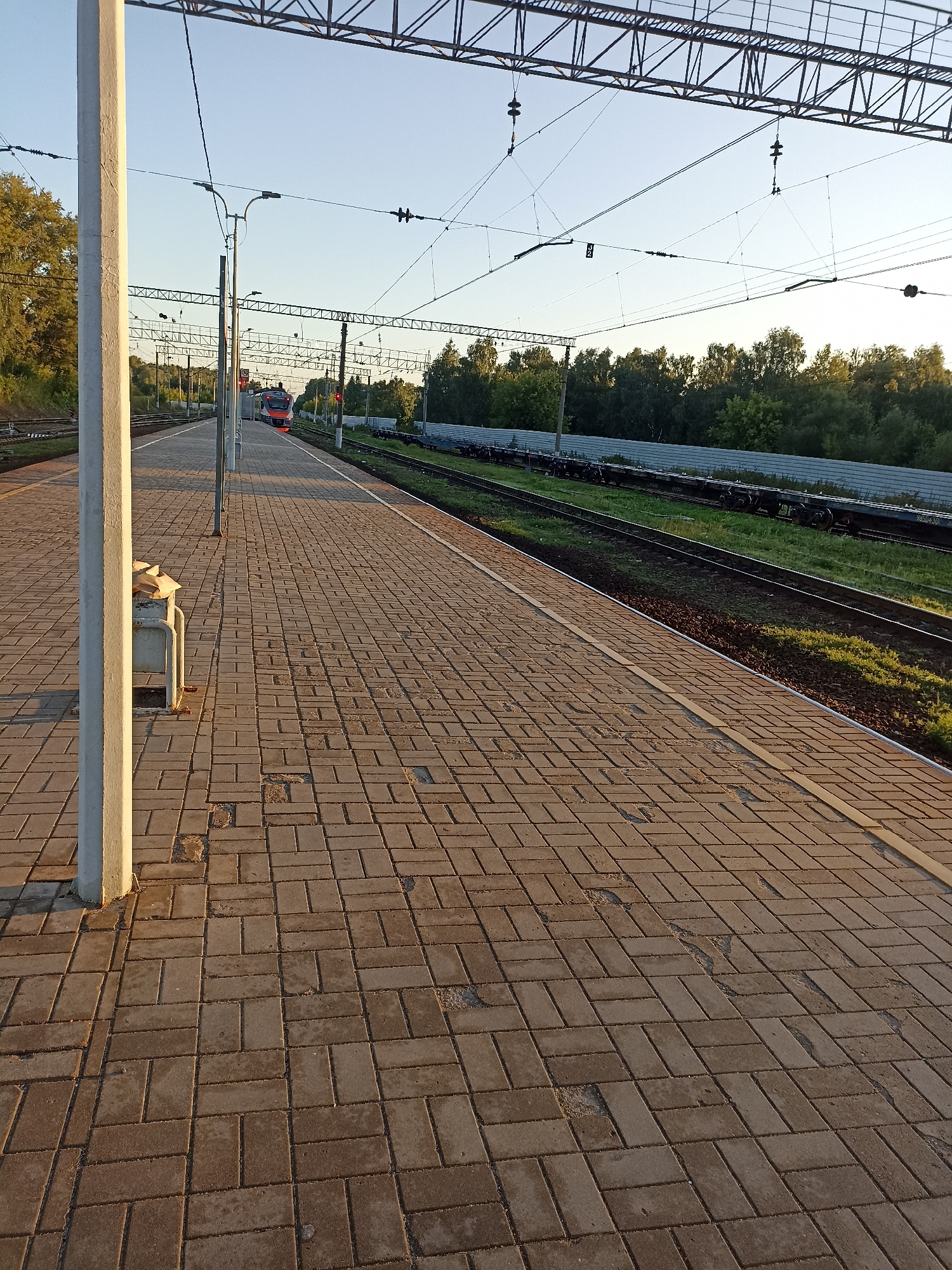 The image depicts a railway platform in Chekhov, Russia, under a clear sky during what appears to be late afternoon or early evening, judging by the low angle of the sun casting long shadows. The platform itself is paved with rectangular bricks arranged in a herringbone pattern, with some areas showing signs of wear and missing bricks. To the left, a tall, light-colored concrete pillar stands prominently, supporting overhead power lines. Beside the pillar, there is a metal bench with a trash receptacle attached, containing some crumpled paper.
In the middle ground, a red and white passenger train is approaching on the tracks. The train has its headlights on. Beyond the tracks, there is a grassy embankment and a line of trees forming a natural border. Further in the distance, a white metal fence runs parallel to the tracks. The overhead structure of power lines, characteristic of electrified railways, is visible across the top of the frame. The overall scene is one of quiet activity at a suburban or regional train station.