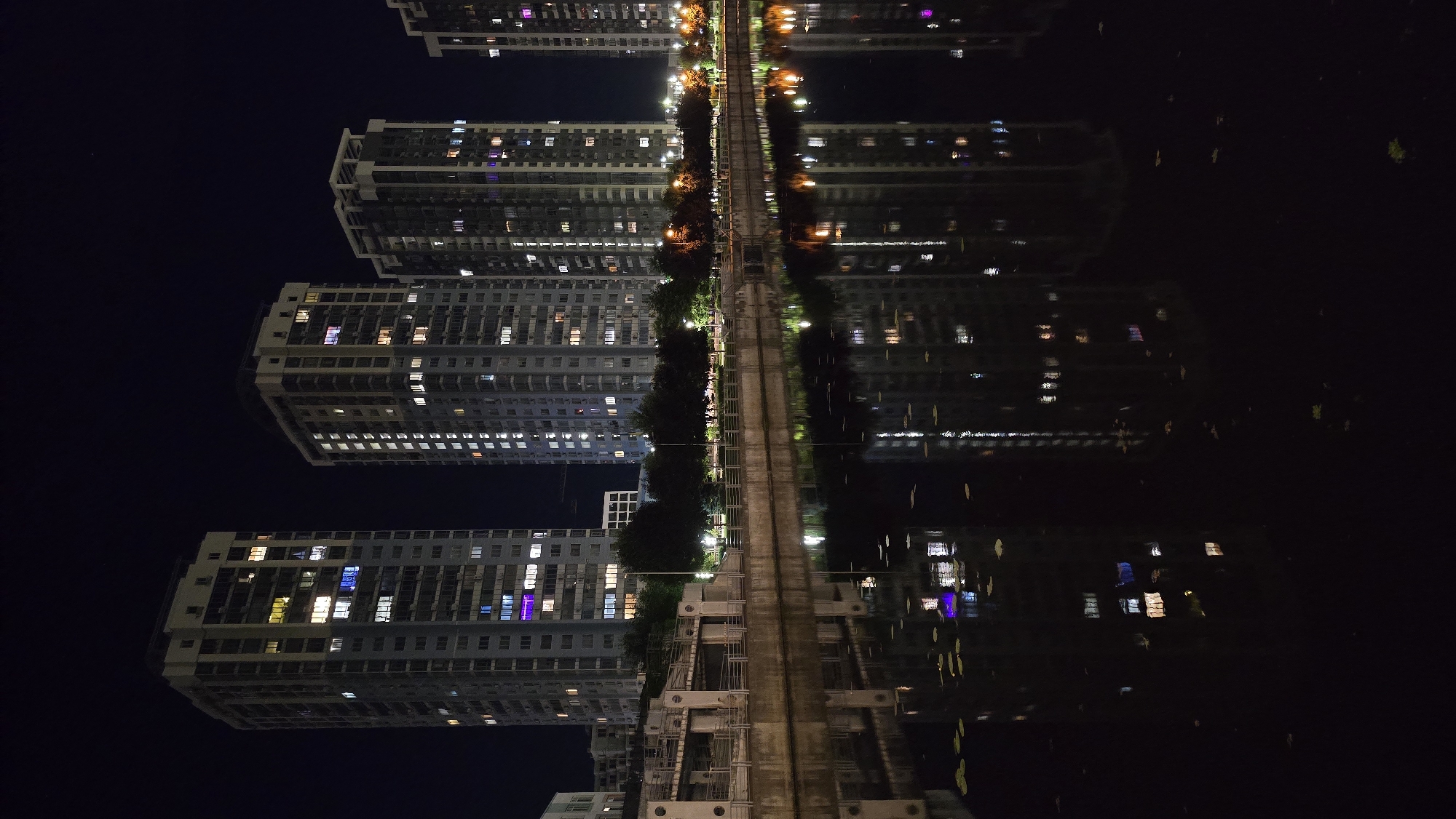 This is a nighttime, vertical, full shot photograph of a body of water with reflections of tall apartment buildings on the opposite bank. The buildings are illuminated from within, with lights visible in many windows, indicating occupancy. The water is dark, with reflections of the buildings, lights, and some scattered leaves or debris. A concrete embankment with a railing separates the buildings from the water. The sky is dark, with no visible celestial bodies. The scene appears to be an urban waterfront area, possibly part of a residential complex. Based on the architecture and the presence of the Dnipro River, this location is likely Kyiv, Ukraine, specifically the residential areas along the river, such as the Obolon district. No people are visible in the photograph.