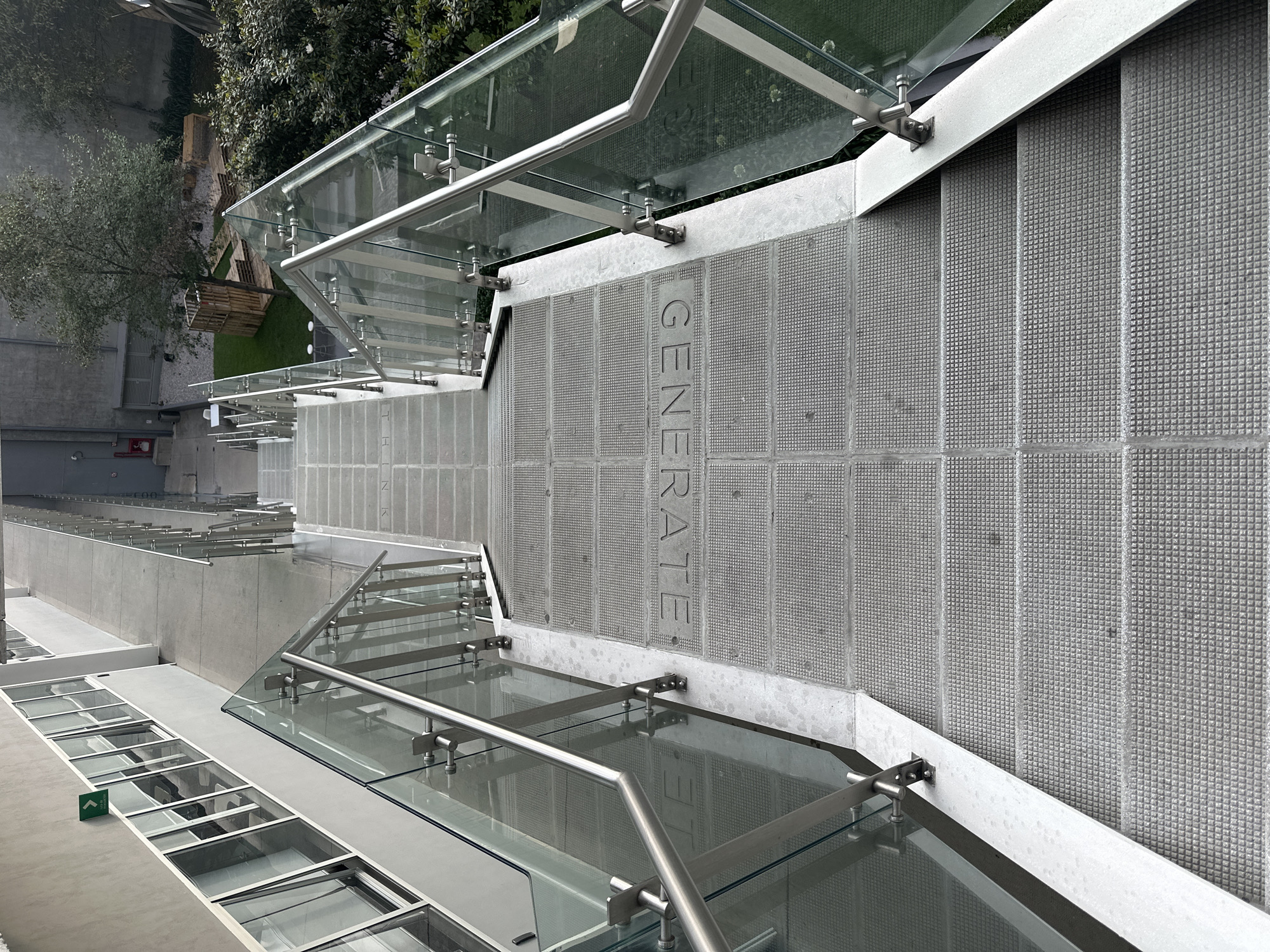The image depicts a downward-angled view of a modern architectural feature, likely stairs or a multi-level structure. The location is in Mexico City, Mexico.
The central element is a set of gray, textured steps. On one side of the steps is a gray wall with the word "GENERATE" vertically inscribed. Above the stairs are glass panels and metal supports, serving as a partial roof or awning. Glass railings with metal supports are present along the edges of the stairs and walkways.
Below the stairs, the image shows a glass-covered walkway or platform, with rectangular glass panels set into a gray frame.
To the upper left, there's greenery, including trees and artificial grass, indicating an outdoor or landscaped area adjacent to the structure. The overall setting suggests a contemporary urban environment, possibly a public space, commercial building, or cultural institution in Mexico City. There are no people visible in the frame.
