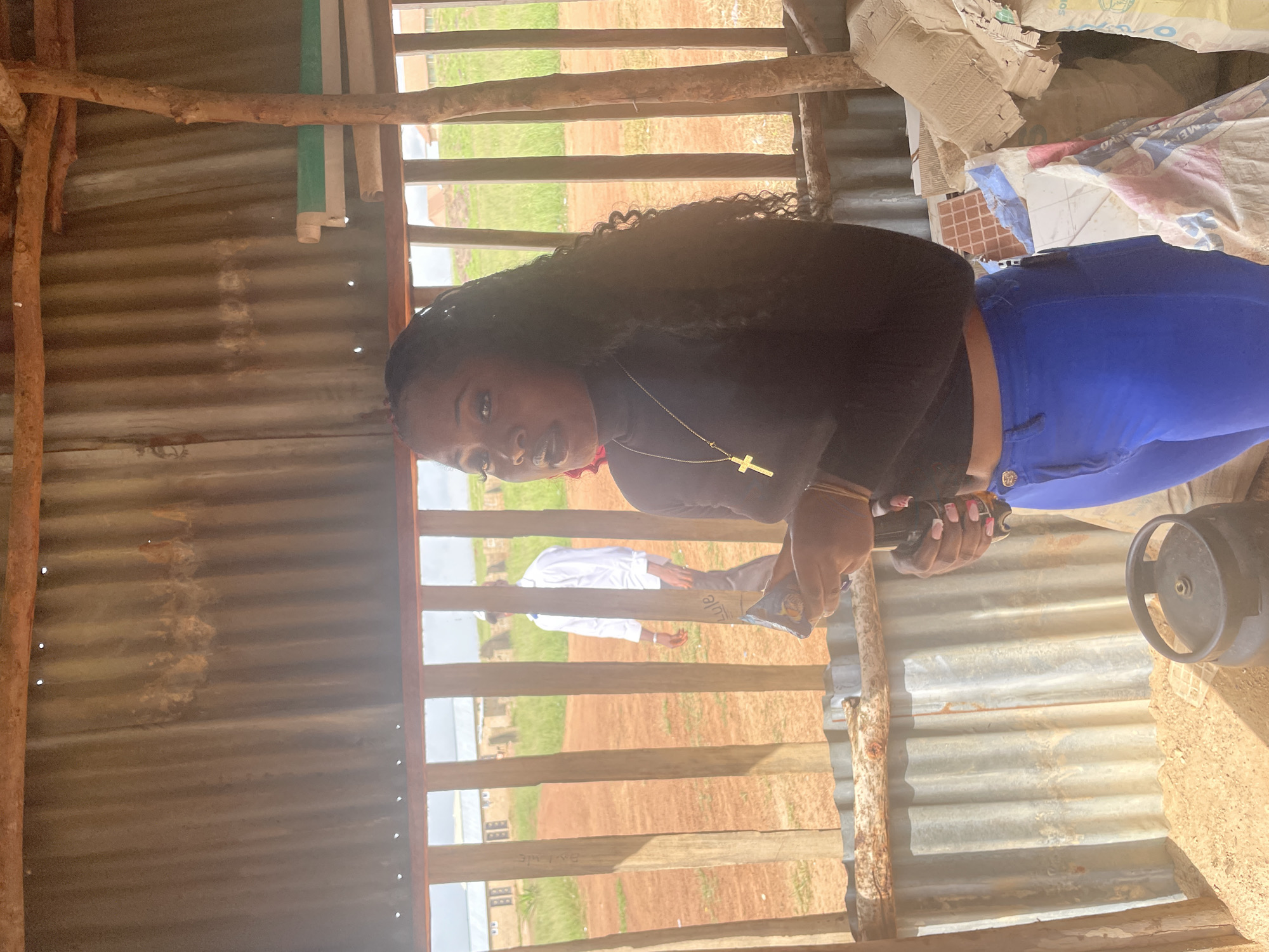 A young Black woman stands in what appears to be an outdoor shelter with corrugated metal walls and a wooden frame. She is wearing a black long-sleeved top and bright blue pants. A gold necklace with a cross pendant is visible around her neck. Her hair is long and dark, styled in curls. Her hands are holding a dark object, possibly a phone or a small device, and her fingernails are painted pink.
In the background, through the wooden slats of the shelter, a grassy, outdoor scene is visible, suggesting a rural or semi-rural setting. The light indicates it is daytime. To the right of the woman, a black gas cylinder with a red top is visible. Some white sacks, possibly containing building materials, are piled up behind her. The overall impression is that of a casual, everyday moment captured within a somewhat rustic environment. The woman's expression is difficult to discern clearly, but she appears to be looking towards the camera. The scene offers no specific text.