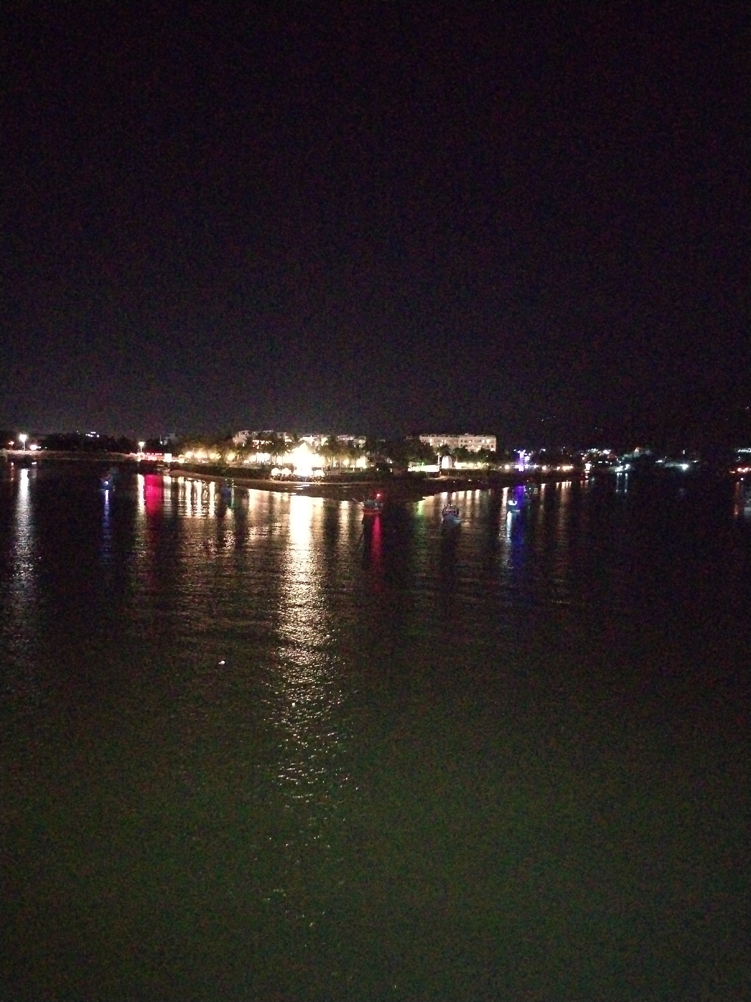 The image depicts a night scene over water, likely a bay or large body of water. Across the water, there is a cityscape with lights illuminating buildings and other structures. The lights are reflected in the water, creating a shimmering effect. There appear to be several boats or small vessels scattered in the water, marked by colored lights. The overall environment suggests a nighttime urban setting with a focus on the waterfront. Given the location in Bac Nha Trang Ward, Vietnam, it is likely that this is the coastal area of Nha Trang.