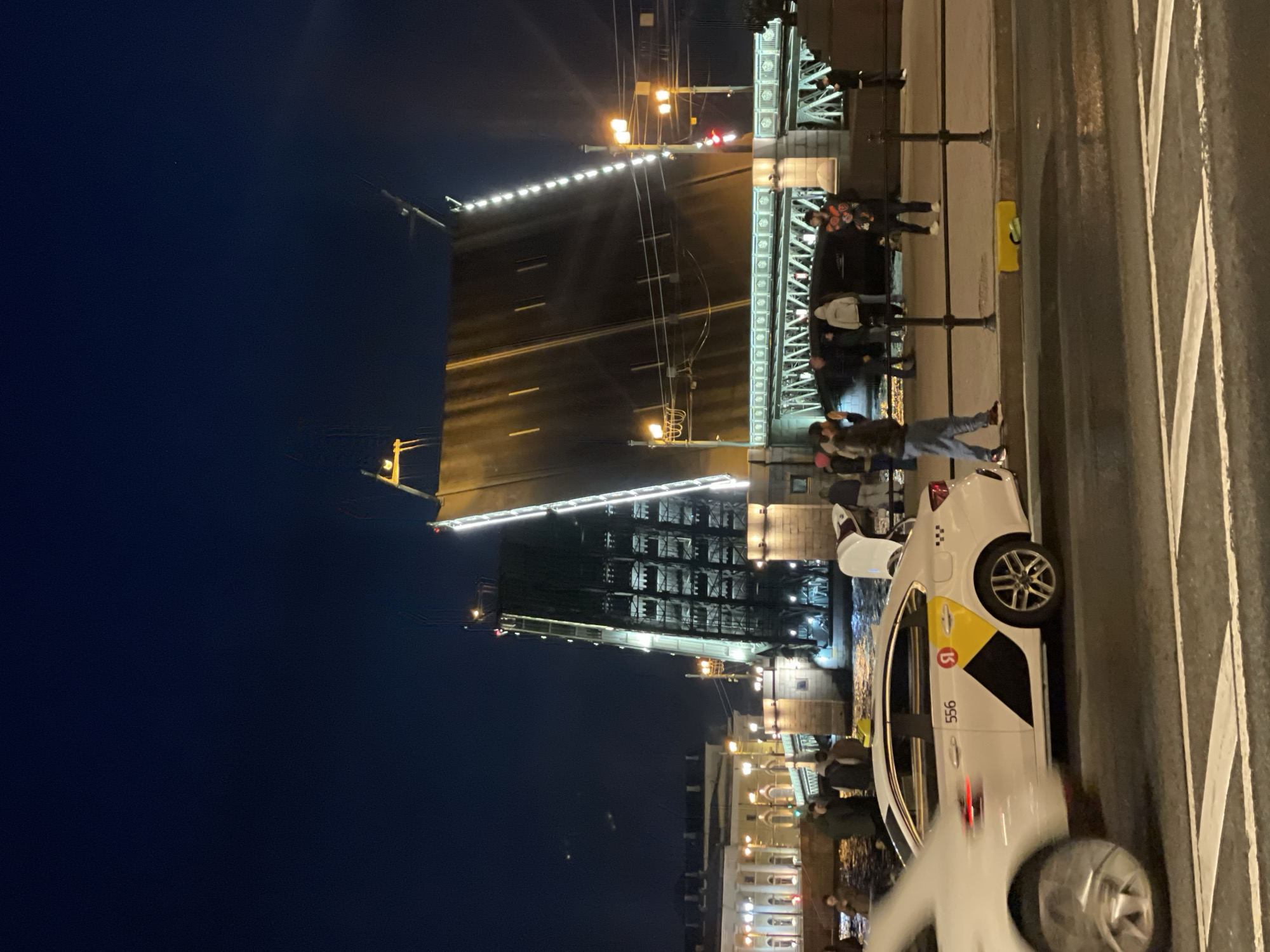 The image captures the Palace Bridge in Saint Petersburg, Russia, during its nighttime opening. The bridge's two sections are raised, allowing river traffic to pass. A crowd of people watches the event from the bridge's edge. A white taxi cab with a distinctive logo is parked on the right side of the frame, partially obscuring the view of the bridge. The sky is dark, contrasting with the bridge's illuminated structure.
