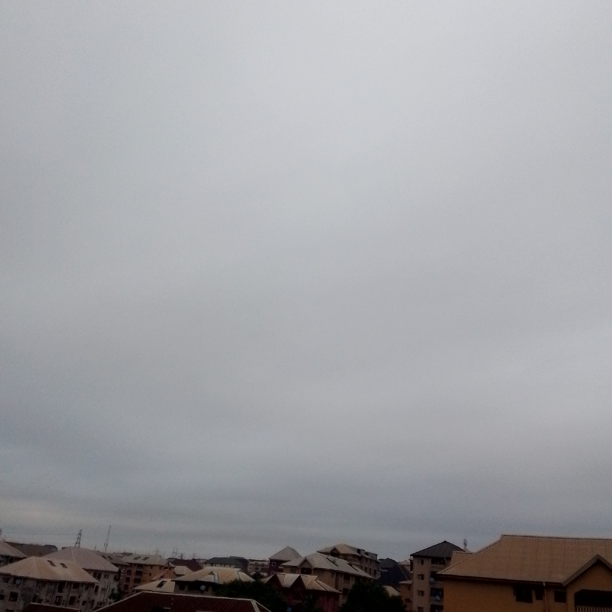 The image shows a cityscape under an overcast sky. In the foreground and midground, multiple buildings with peaked roofs are visible, constructed with various materials like brick and possibly painted surfaces. Some of the buildings appear to be residential, with multiple stories and windows. A few utility poles and antennas are also present in the scene. The overall atmosphere is subdued, with muted colors due to the cloudy weather. The location is identified as Unknown, Unknown.