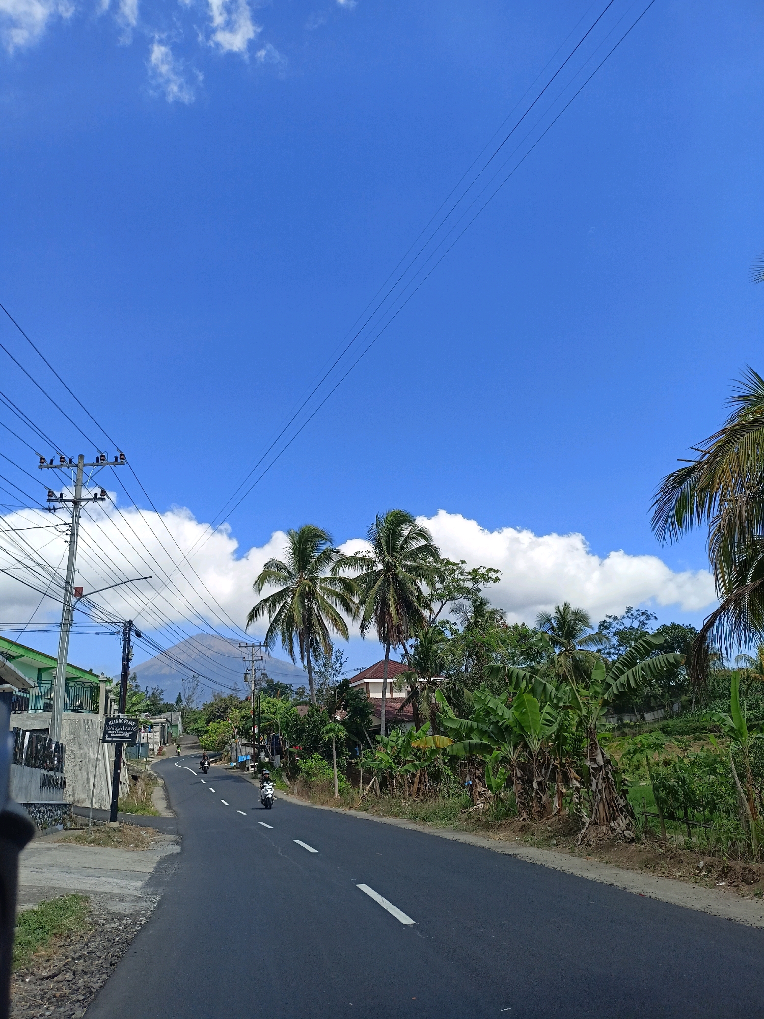 A rural road in Windusari, Indonesia, winds through a lush landscape under a bright blue sky with scattered white clouds. On the left side of the road, a building with a green roof and a concrete wall is visible, along with a utility pole adorned with numerous wires. Further down the road, two motorcycles are being ridden. The right side of the road is lined with banana trees and other tropical vegetation. In the distance, a tall, conical mountain is silhouetted against the sky, possibly Mount Merapi. The road is paved and has white dashed lines indicating lanes. The overall scene depicts a tranquil, everyday scene in a tropical rural setting.