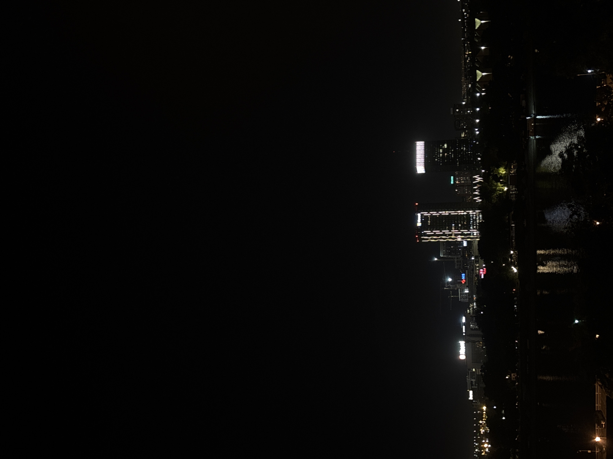 This is a nighttime shot of the skyline in Belgrade, Serbia, taken from across a body of water, likely the Sava River. The city lights of modern high-rise buildings are visible, reflecting on the surface of the water. The silhouette of trees along the riverbank can also be seen. Streetlights are distributed along the riverside.
