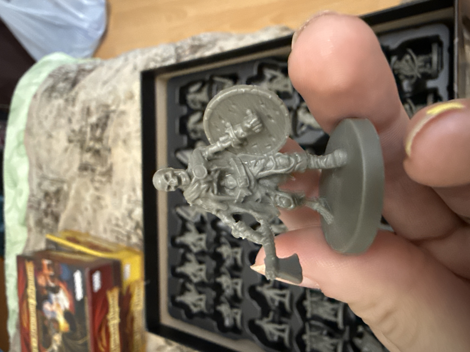 The image shows a person holding a gray plastic miniature of a skeleton warrior in their hand. The miniature is armed with a shield and an axe, and is mounted on a round base. The background shows a black plastic tray filled with identical skeleton warrior miniatures. In the lower left corner, partially visible, are boxes of a board game. The setting appears to be indoors. There is no evidence in the image to suggest the location is Moscow, Russia.