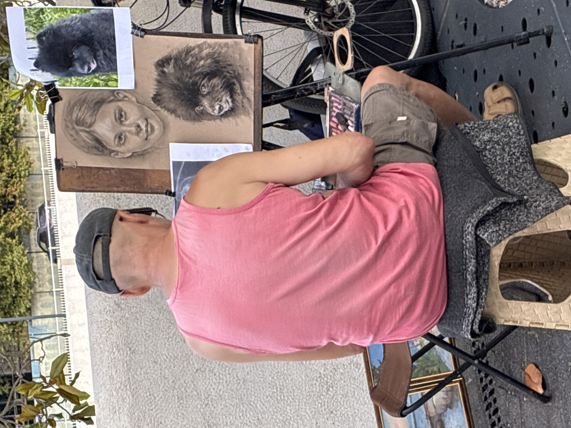 A man wearing a pink tank top and a grey baseball cap is sitting on a stool in front of an easel in Sochi, Russia. He is bald and has sunglasses resting on his head. The easel displays two charcoal drawings: one of a young girl and another of a black dog. A reference photo of a black dog is clipped to the easel above the drawings. The man appears to be in the process of painting or drawing. In the background, a bicycle is visible, as well as a white fence and some greenery. The ground is paved with grey stones and there is a folded chair with a framed painting on it to the left of the man.