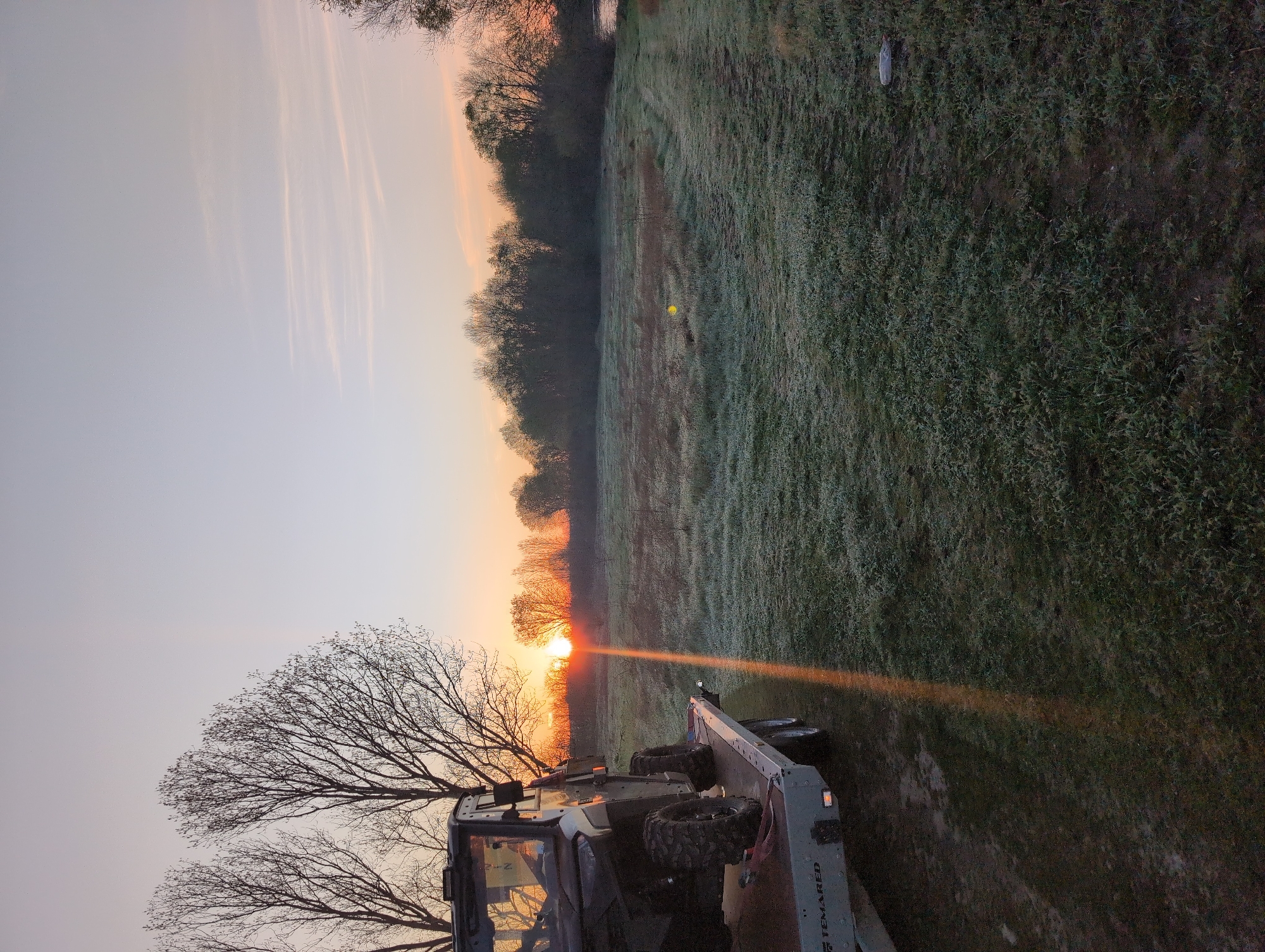 The image depicts a sunrise scene near Zetiovo, Bulgaria, showing a field covered in frost. The sun is rising on the horizon, partially obscured by trees. A utility terrain vehicle (UTV) is parked in the foreground. The sky is a gradient of colors from pale orange to light blue. The field has a yellow golf ball on the frosted grass.