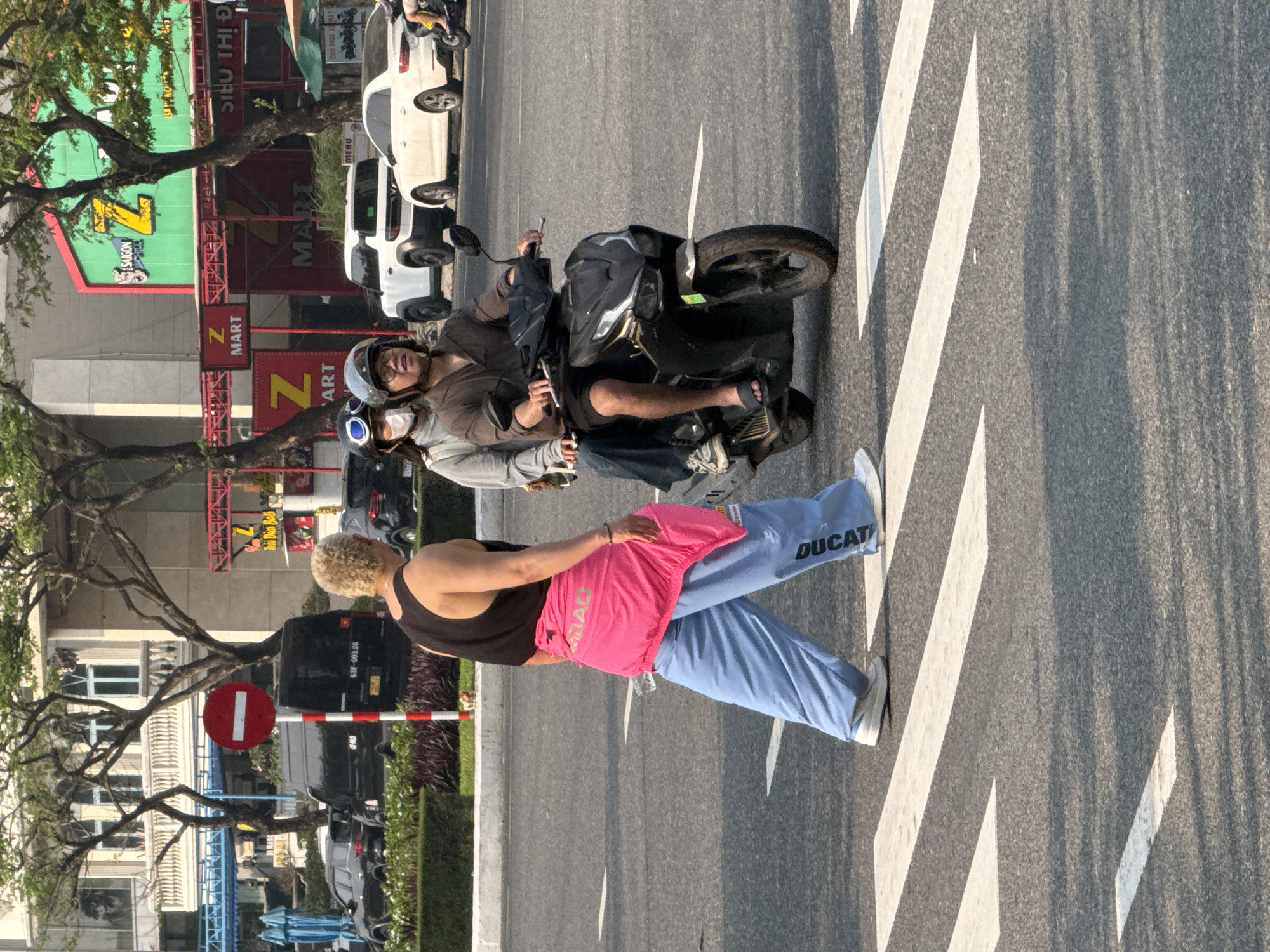 A man drives a motorbike through a crosswalk in a city in Vietnam, with a female passenger on the back. The driver is wearing a brown shirt, dark pants, a gray helmet, and a white face mask. He is wearing open-toed sandals on his feet. The motorbike is dark colored with a bag strapped to the back of the seat. The woman passenger is wearing a black tank top, a pink garment wrapped around her waist, and blue sweatpants with the word "DUCATI" down one leg. She is wearing white sneakers. The crosswalk is made of white stripes. The street is asphalt. Behind the crosswalk is a low barrier with a red-and-white striped pole and a no-entry sign on top. Beyond the barrier is a row of parked cars. Across the street are some businesses, including Z-Mart and Saigon Mart. Trees are interspersed with the buildings. Street signs in Vietnamese are visible. The image appears to be taken in Đà Nẵng, Vietnam.