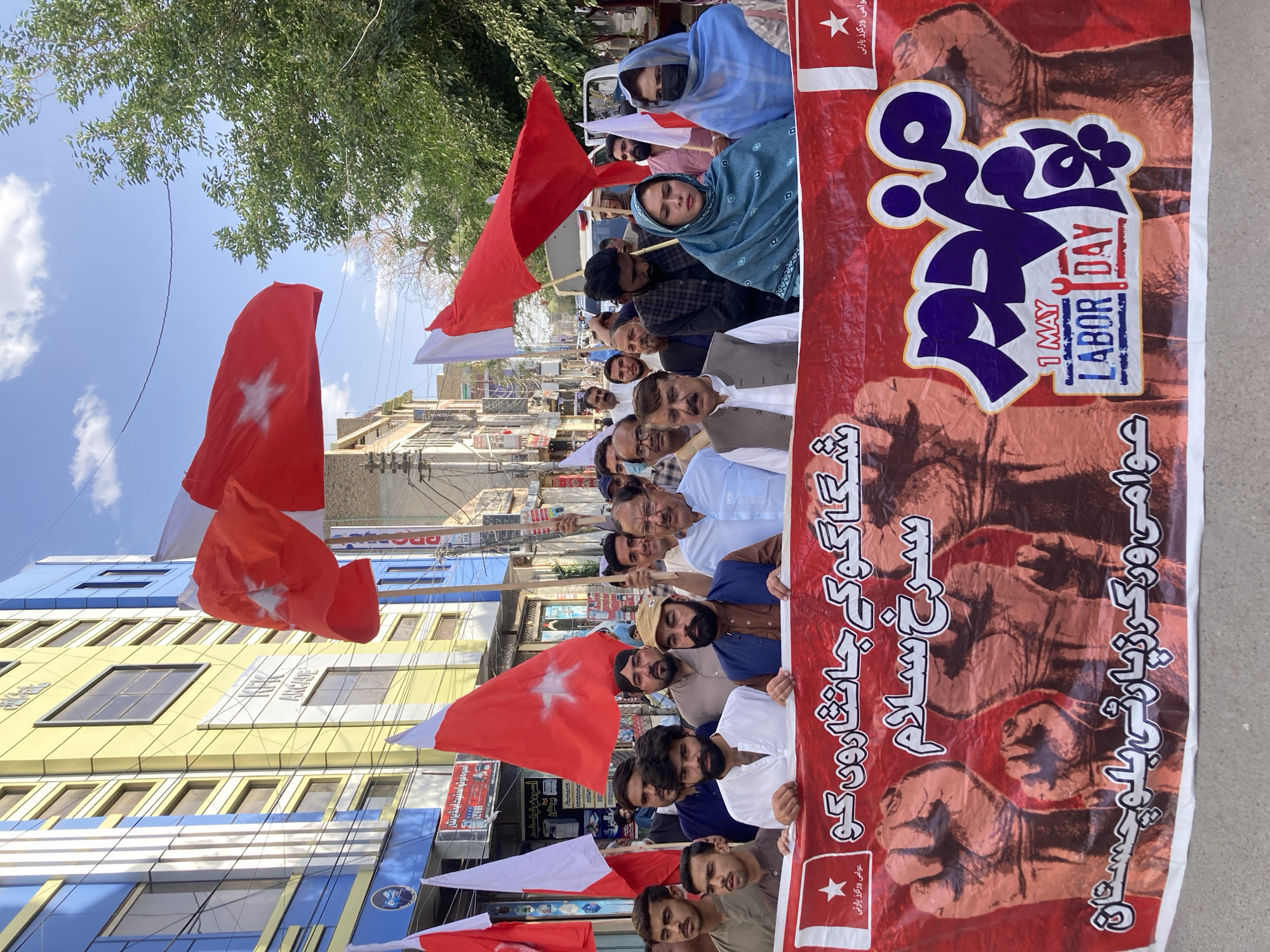 The image depicts a political rally in Quetta City Tehsil, Pakistan. A diverse group of men and women are marching, holding red and white flags with a star symbol, and carrying a large red banner. The banner commemorates May 1st, Labor Day, with text in Urdu and English. The backdrop features multi-story buildings with shops on the ground floor.