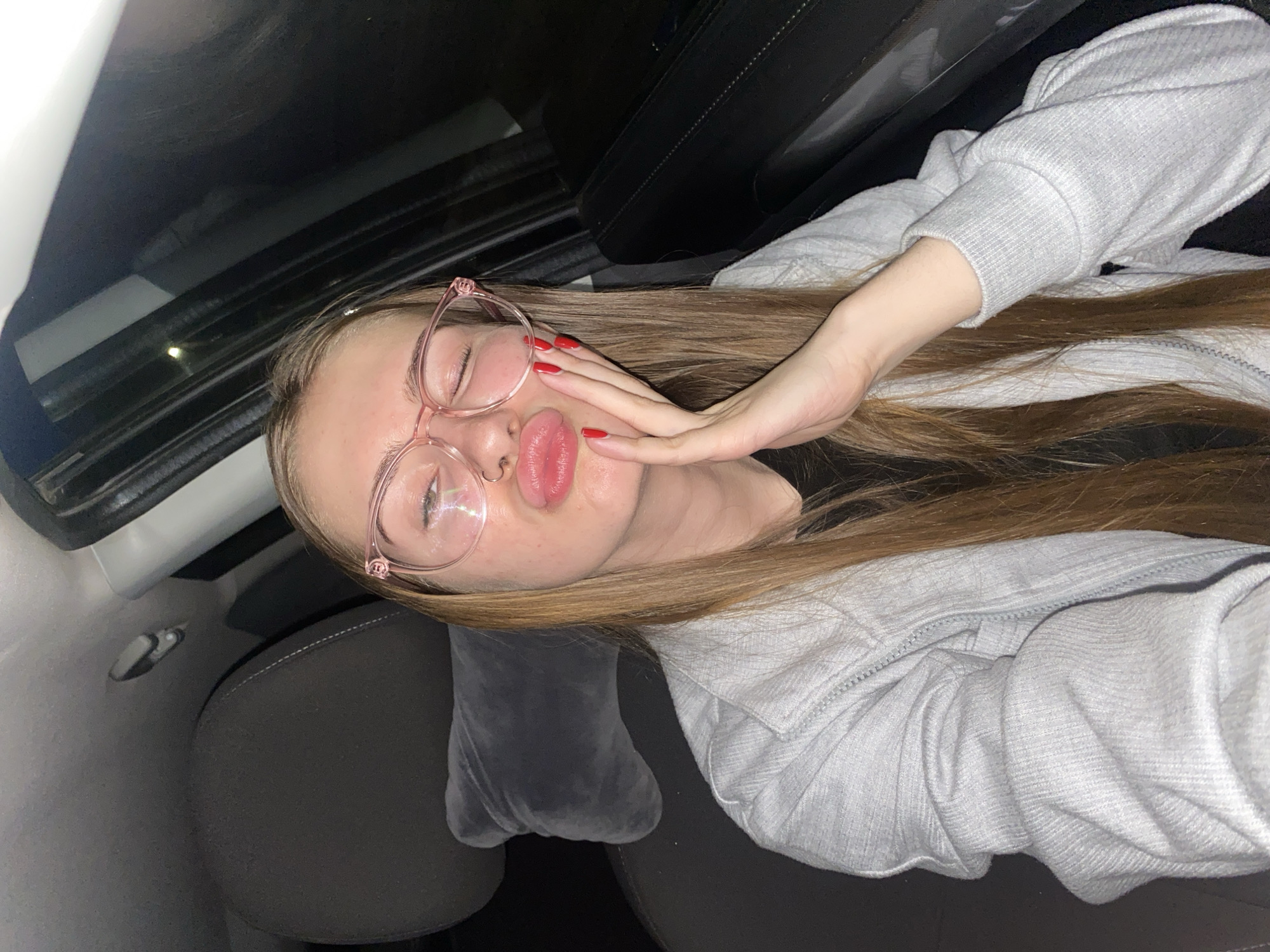 A young Caucasian woman is pictured inside a car, appearing to be in the city of Dyakovo, Russia. She is wearing clear-rimmed pink glasses and a light gray hooded jacket. Her eyes are closed and her lips are pursed in a kissing gesture. Her right hand is placed on her cheek, with bright red nail polish visible on her fingers. The car's interior, including a dark gray headrest and part of the seat, is visible. The lighting suggests it is nighttime or a dimly lit environment.