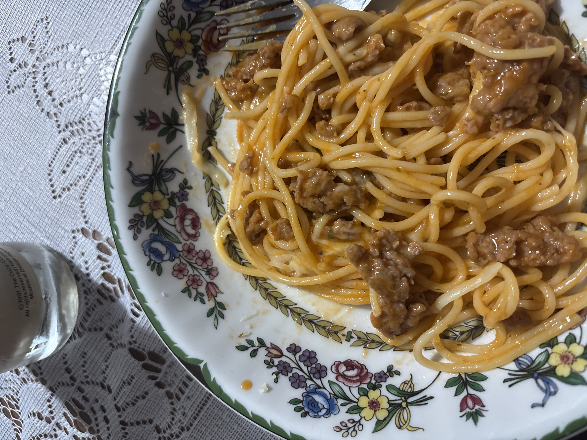 The image shows a plate of spaghetti with meat sauce, topped with a fork, sitting on a white lace tablecloth. A partial view of a glass bottle is visible to the left of the plate. The plate has a floral pattern with a green trim. The location is not depicted in the image and therefore cannot be determined.