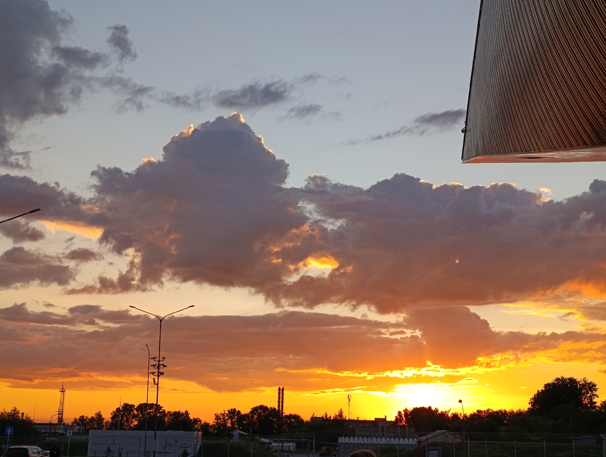 This media file depicts a dramatic sunset or sunrise over an urban or industrial fringe landscape in Bolshoye Savino, Russia. The sky is the dominant subject, filled with a dynamic display of colors and clouds. The upper portion of the sky is a pale blue, gradually transitioning to vibrant oranges and yellows near the horizon, suggesting the golden hour.
Numerous clouds are scattered across the sky; some are large and dark grey, while others have bright, glowing edges where they are illuminated by the unseen sun. A particularly prominent, towering cloud mass is located in the upper-mid section, also featuring a radiant, sun-kissed top.
Silhouetted against the brilliant sky are various features of the landscape below. Dark, leafy trees line the horizon, interspersed with industrial-looking structures such as tall towers or chimneys. Two distinct streetlights or lampposts stand in the mid-ground, their intricate forms starkly outlined by the intense light behind them. Hints of buildings or low structures are visible at the very bottom.
A notable detail is a large, dark, angled structure in the upper right corner, with a textured, vertically striped surface. It appears to be an architectural element, possibly an overhang or part of a building, extending into the frame and partially obscuring the sky. No discernible text is visible. The overall mood is one of natural beauty and urban melancholy, characteristic of an evening sky.