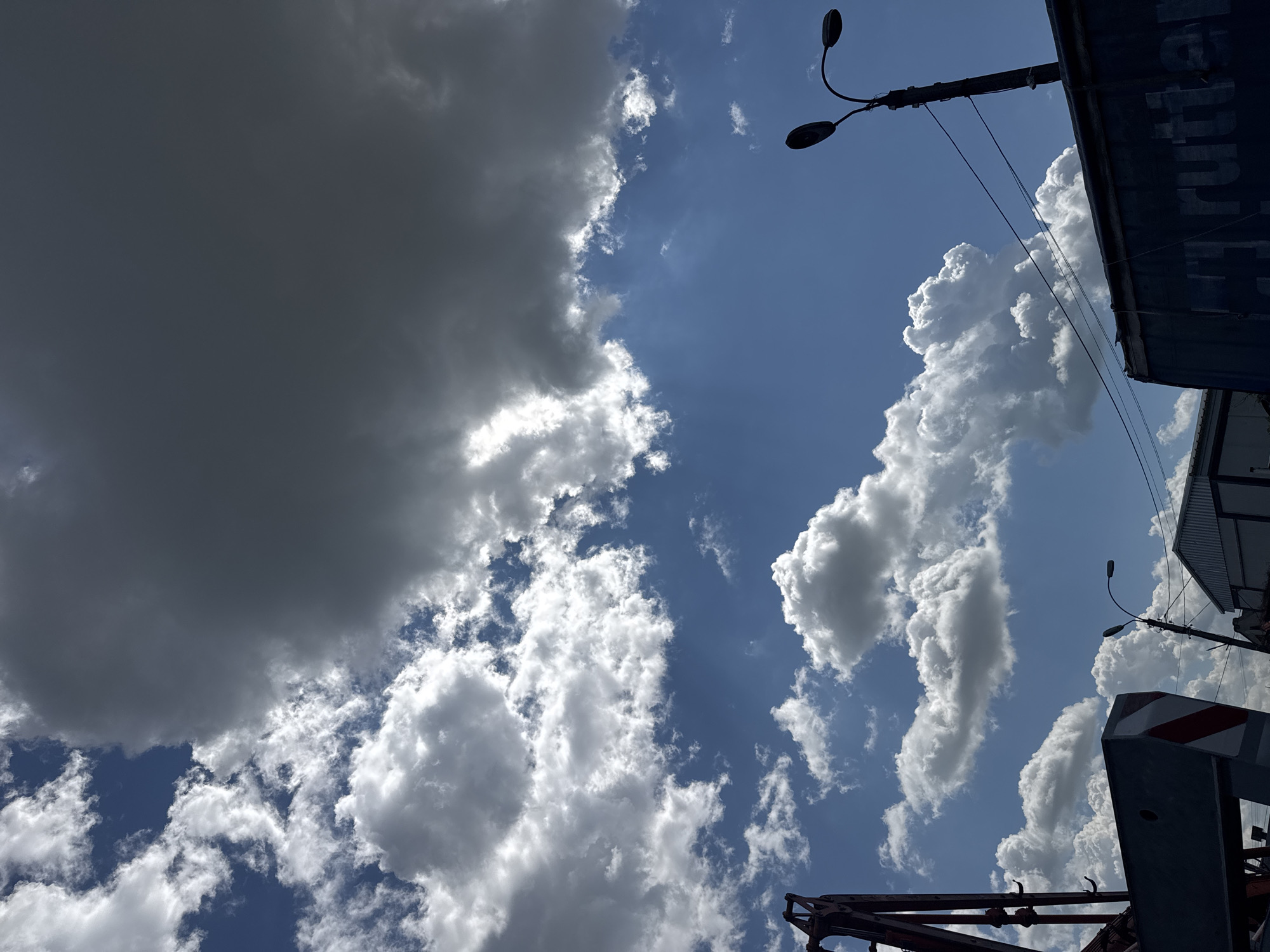 A low-angle shot captures a dynamic sky filled with contrasting clouds over an industrial area. In the upper left, a dark, ominous cloud bank looms, partially obscuring the bright blue sky and casting a shadow. Scattered white, fluffy cumulus clouds dot the lower half of the sky, illuminated by sunlight. In the foreground, to the left, is a portion of a red construction crane. Below the crane and to the right, a blue shipping container is visible, with partial lettering that reads "y rutter". A street light with two lamps stands to the right of the container, with its wires extending across the frame. In the background, several industrial buildings with corrugated metal roofs are discernible. The overall scene suggests a partly cloudy day with signs of ongoing construction or industrial activity.