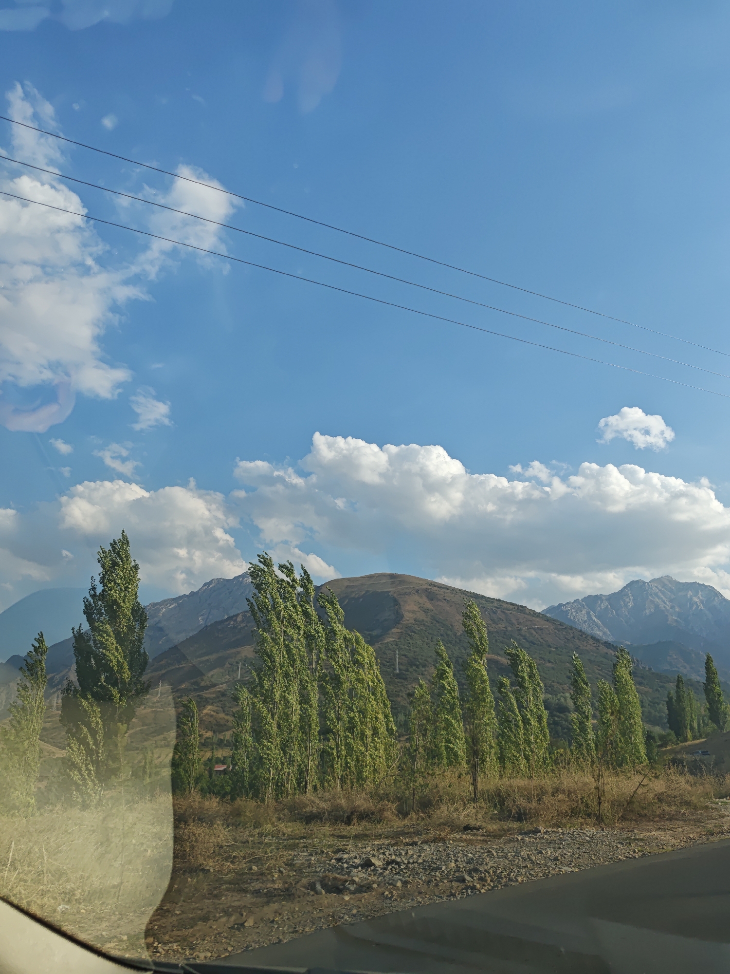 The image depicts a landscape scene with a clear blue sky dotted with white clouds. Several utility wires stretch horizontally across the upper portion of the frame. In the foreground, a road is visible, with a section of what appears to be a car's windshield reflected on the left side. Beyond the road, a line of tall, slender trees with green foliage stands against a backdrop of rolling hills and mountains. The terrain is dry and appears to be covered in sparse vegetation and rocks. A hint of a building with a red roof is visible between the trees in the middle ground. The overall impression is of a rural or semi-rural environment, possibly captured from a moving vehicle. The specific location in the city of Yangikurgan, Uzbekistan, is not explicitly indicated by any visible landmarks or text within the image.