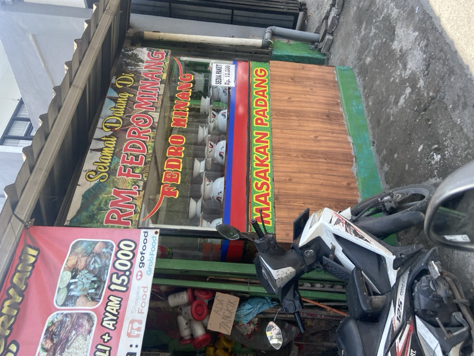 The image shows a storefront in Kecamatan Dayeuhkolot, Indonesia, with a sign that reads "Selamat Datang Di RM. FEDRO MINANG" in red letters on a yellow background. Below the sign is another, smaller sign that reads "FEDRO MINANG" in black letters on a white background. Below this, the window displays various bowls and plates, and the sign reads "MASAKAN PADANG" in green and yellow letters on a red background. A white motorcycle is parked in front of the store. The storefront has wood paneling and is partially obscured by a green wall. The scene is taken from the perspective of a person standing near the motorcycle, looking towards the storefront. The image captures a moment in the daily life of a small business in Indonesia.
