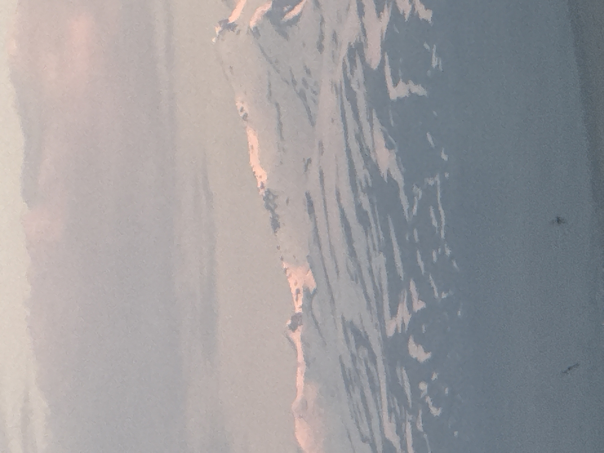This is a close-up, horizontal, medium-shot photograph of a snow-capped mountain range under a pale, pinkish-gray sky. The sky occupies the upper third of the frame, with subtle gradients of color suggesting a sunset or sunrise. The mountain range fills the middle and lower portions of the image. The peaks are covered in snow, with patches of darker rock or shadow visible in between. Streaks of pink light from the sky are reflected on the snow-covered slopes, particularly along the ridgelines. The lower parts of the mountains appear to be in shadow, with a bluish-gray hue. In the very bottom of the frame, there are two small, dark, indistinct shapes that appear to be birds in flight. The overall impression is of a serene, natural landscape. This mountain range is located in the vicinity of Abovyan, Armenia.