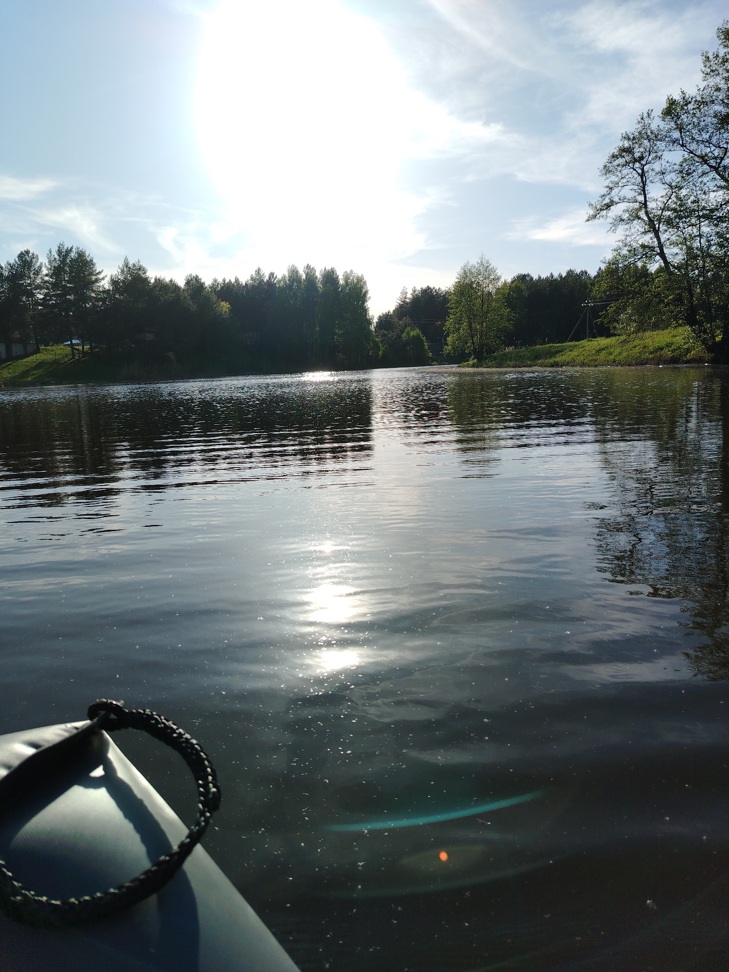 The image captures a bright, sunny scene from an inflatable boat on a calm body of water, likely a lake or river, in Stroitel', Russia.
In the foreground, the front portion of a dark-colored inflatable boat, possibly a kayak or raft, occupies the bottom left corner. A black braided rope is prominently visible on its bow.
The expansive surface of the water dominates the lower and middle sections of the image. It is calm with gentle ripples, and the intensely bright sun creates a strong, shimmering reflection stretching across its surface towards the viewer. Numerous small specks or particles are visible on and just beneath the water's surface, particularly around the boat. Faintly visible beneath the surface in the bottom right are a subtle turquoise shape and an orange circular reflection, possibly indicating submerged objects or optical artifacts.
The background features the far bank of the water body, which is lined with a dense forest of evergreen and deciduous trees, appearing lush and green. The trees cast dark reflections onto the water. On the left side of the shoreline, partially obscured by trees, are hints of small buildings or houses. On the right, the bank appears grassy and gently sloping, with what looks like a power pole or utility structure visible amidst the trees.
The sky above is bright and mostly clear, with some thin, wispy white clouds. The sun, high in the sky, is exceptionally bright and causes significant lens flare, creating a strong overexposed area at the top center of the image.
The scene conveys a peaceful and pleasant daytime setting, likely late morning or early afternoon due to the high sun. No people are visible in the image. There is no visible text. The overall impression is one of natural beauty and tranquility, suggesting a leisurely boat ride.